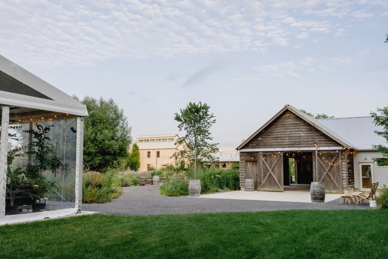 Rustic farm event venue with glass marquee tent, wooden barn, and manicured garden path.