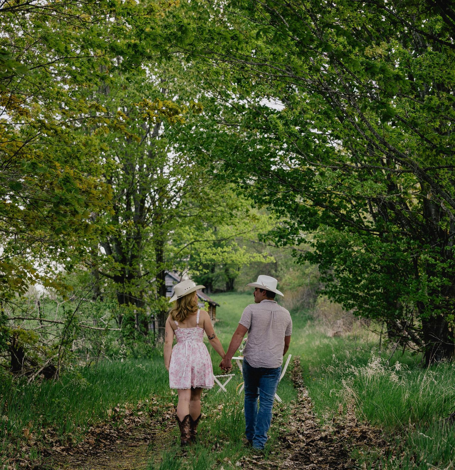 Couple holding hands walking down a scenic tree-lined dirt path wearing matching cowboy hats.