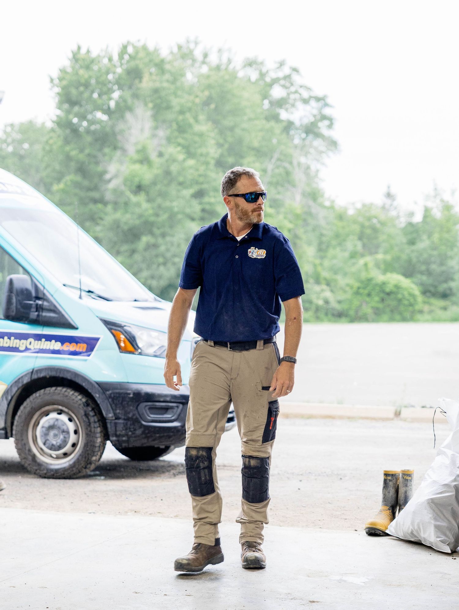 Technician in navy polo shirt walking near a branded plumbing service van in a parking lot.