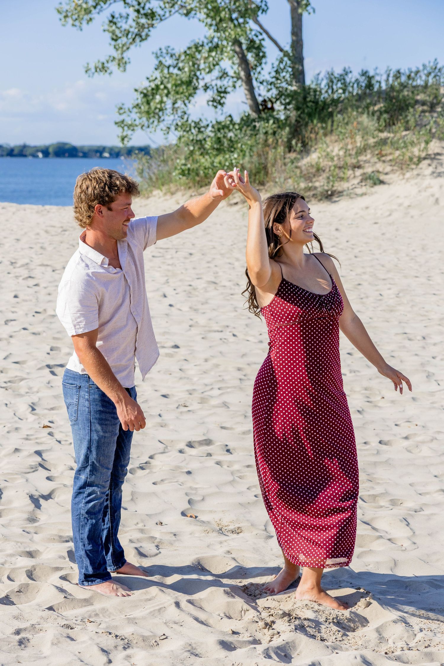 Couple dancing on sandy beach near water, man in white shirt spinning woman in red polka dot dress.