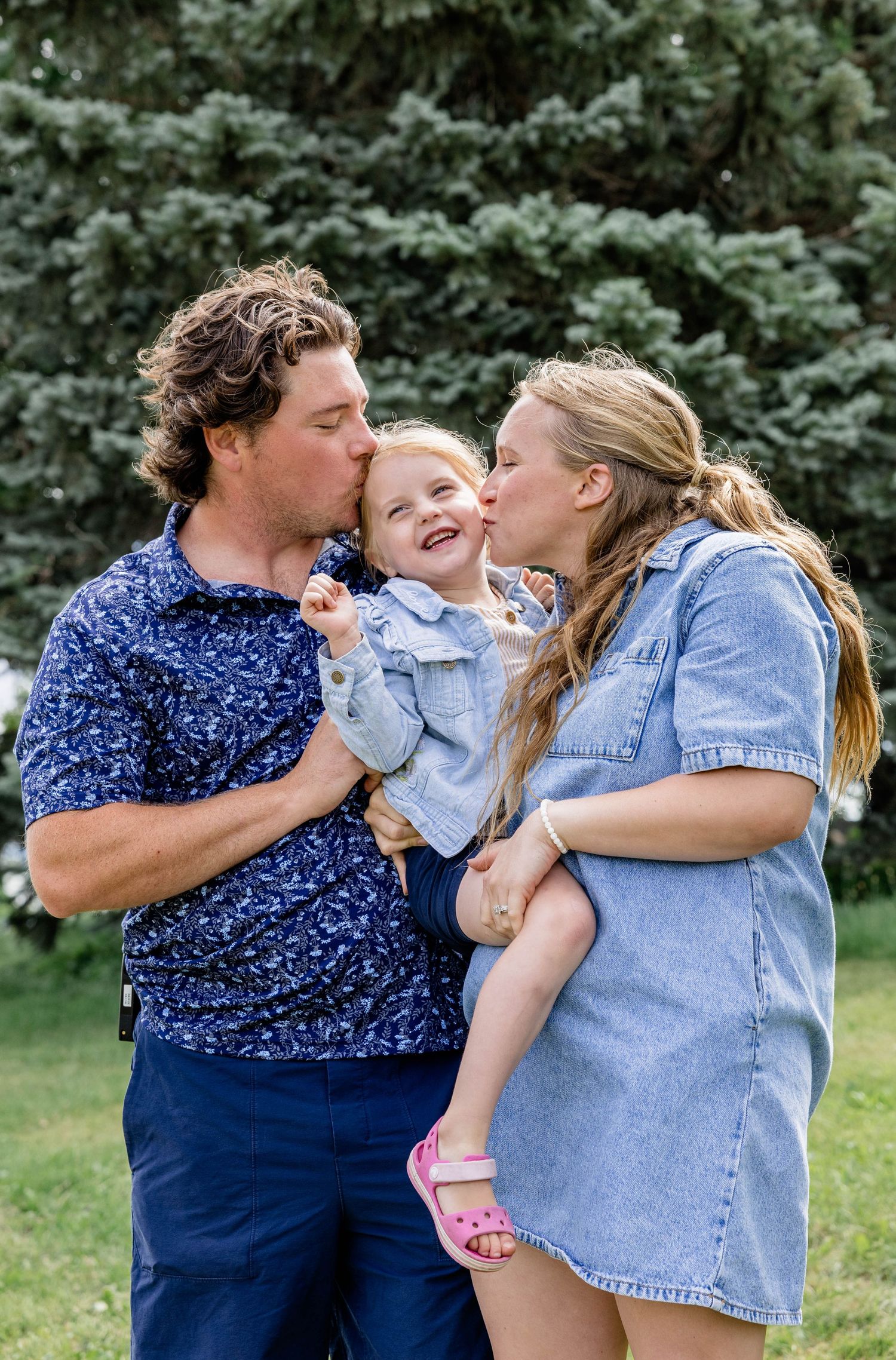A couple kisses their smiling toddler on the cheeks outdoors, with lush green trees in the background.