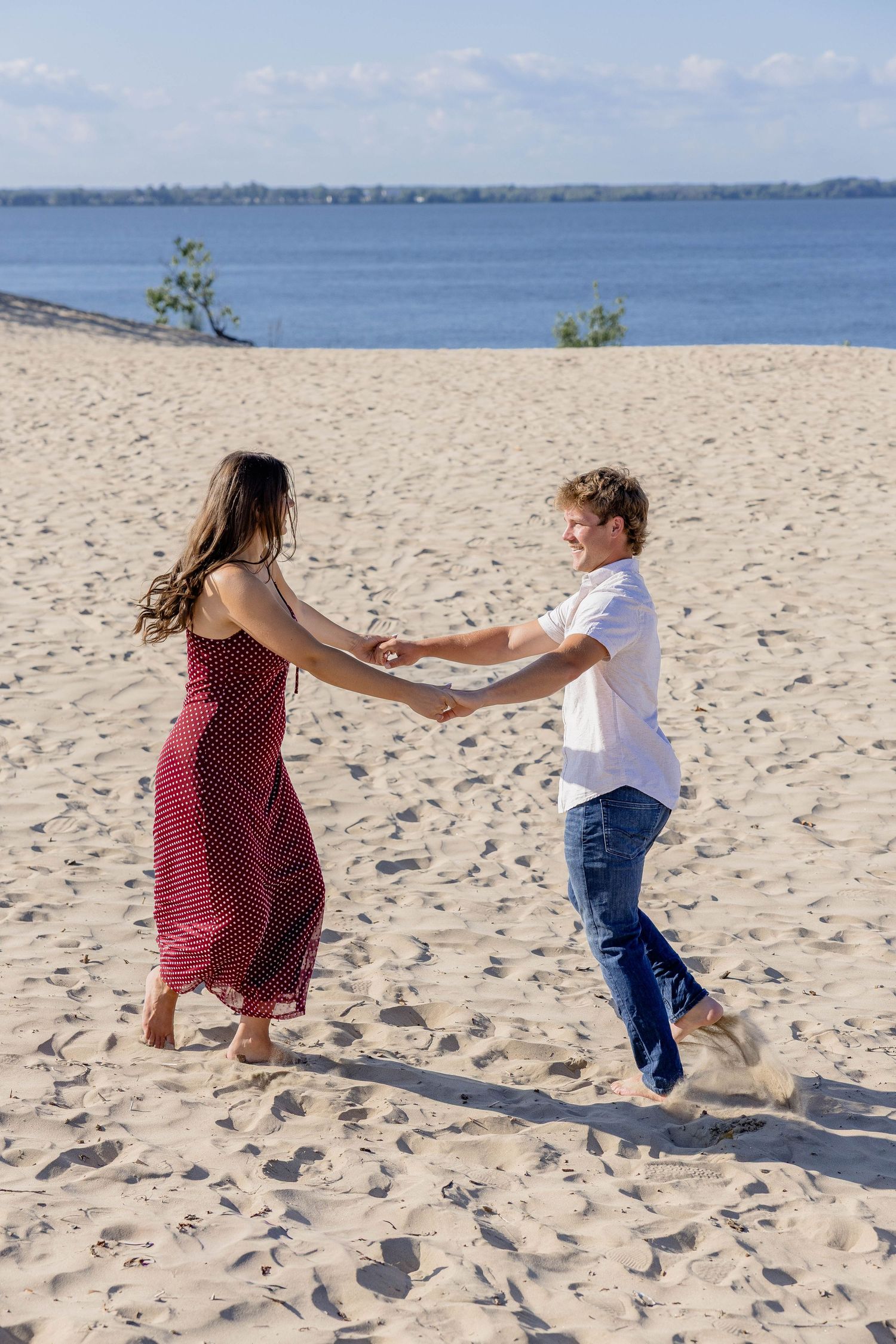 A couple dances joyfully on a sandy beach, holding hands, with blue water and sky in the background.