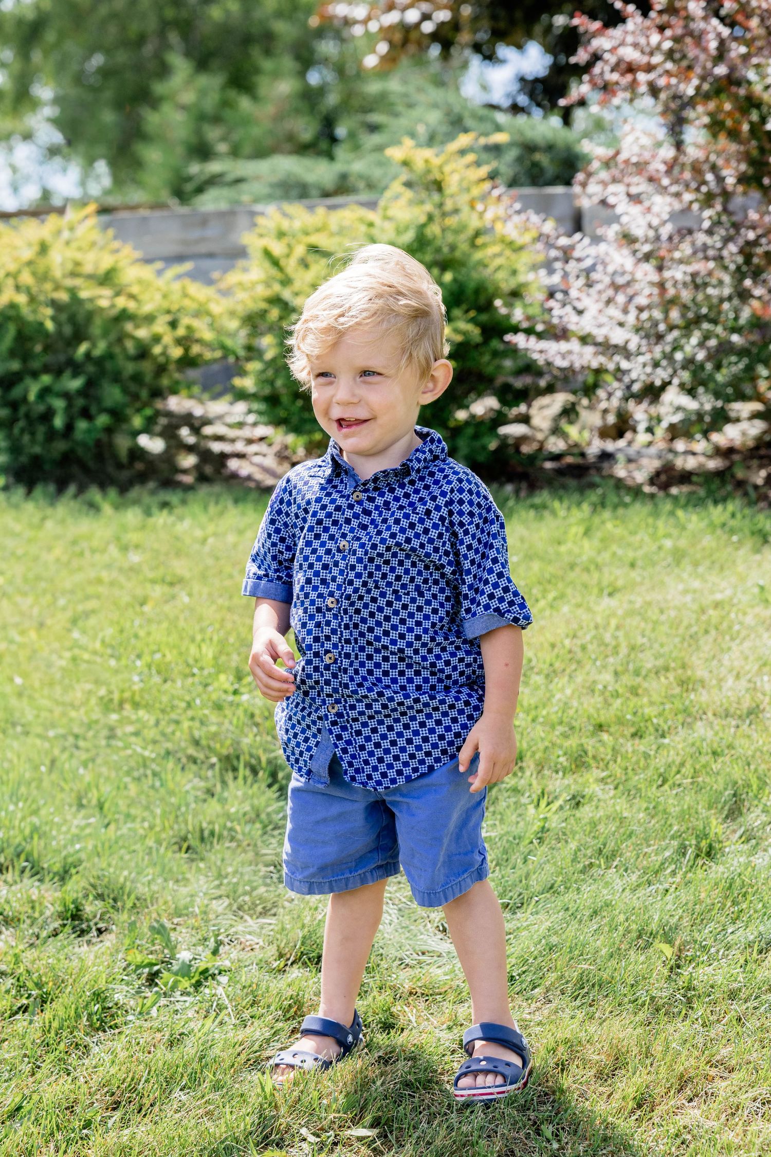 Smiling toddler boy in blue polka dot shirt and denim shorts standing on green grass in sunny garden.