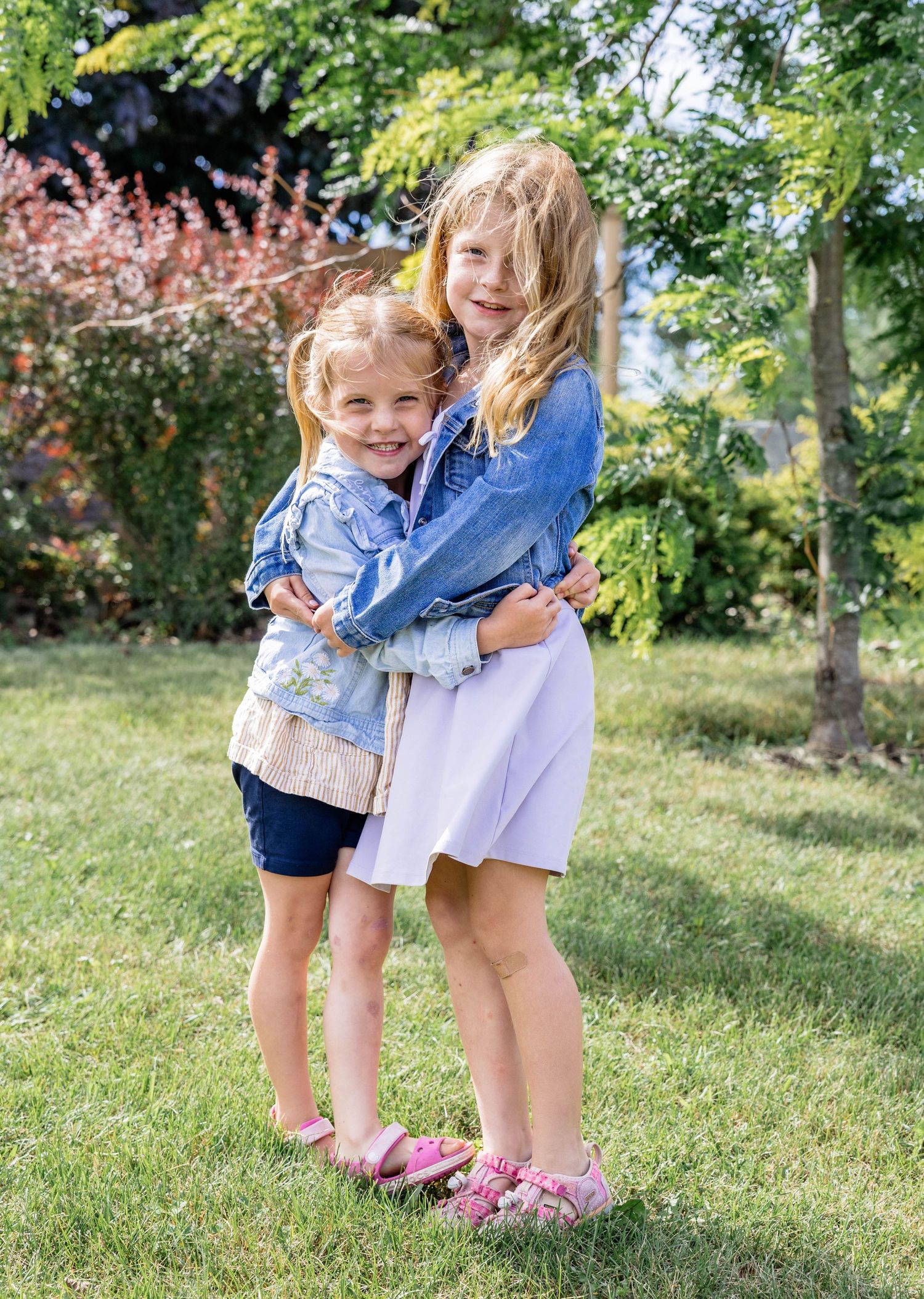 Two young girls in denim jackets hugging and smiling outdoors on a sunny day in a green garden.
