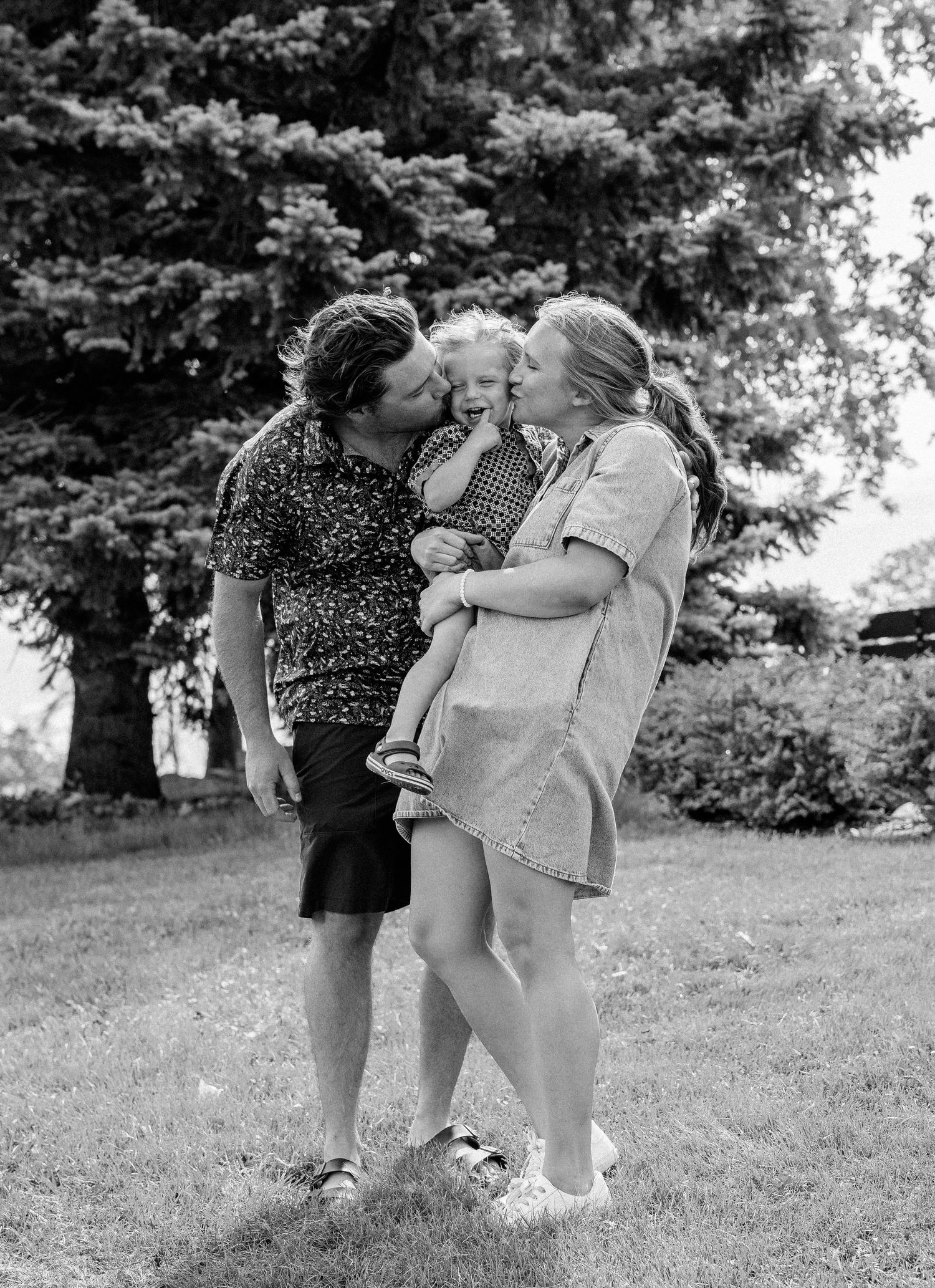 Black and white photo of a couple kissing their toddler outdoors on a sunny day in a lush green yard.