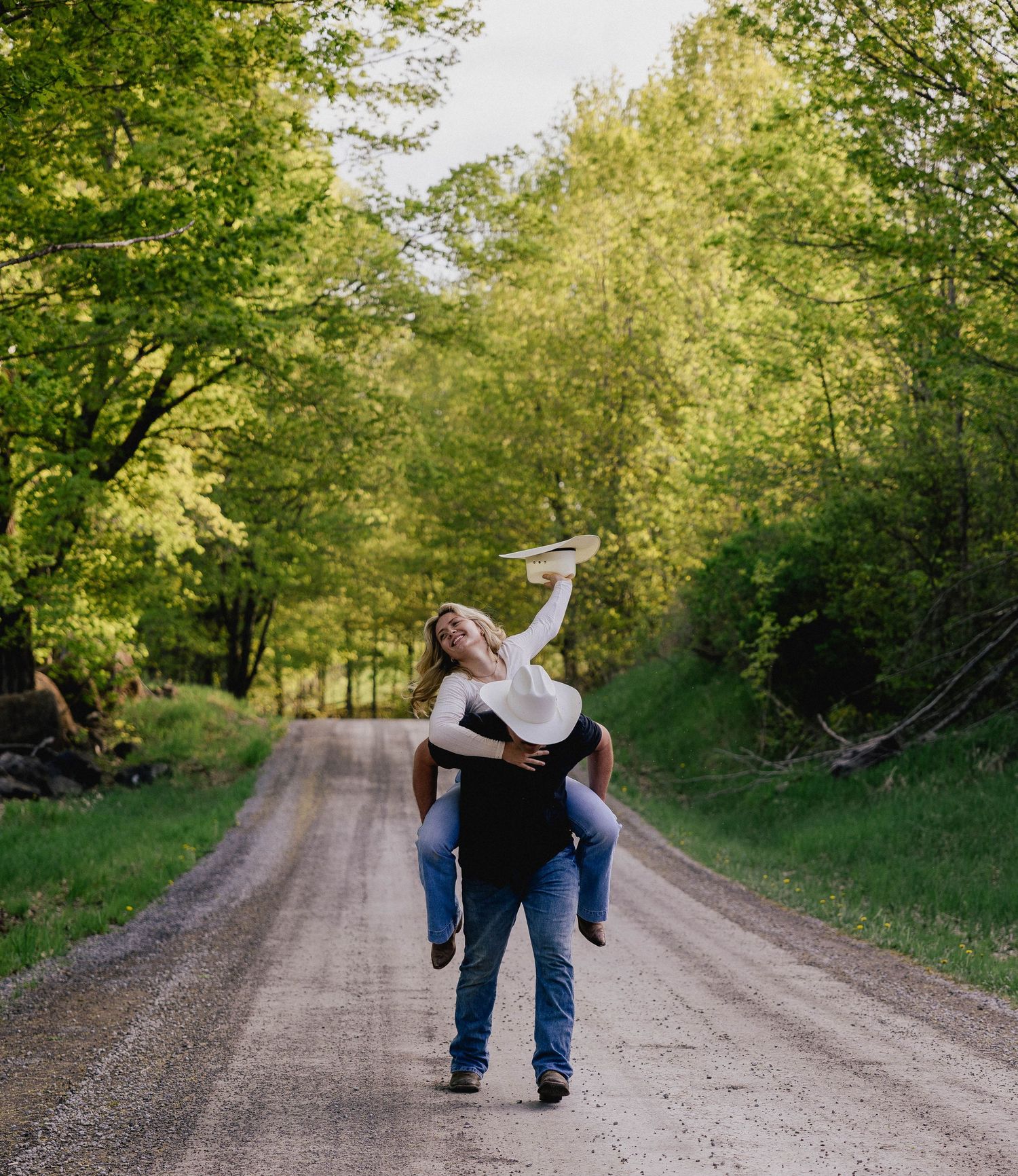 Man giving woman piggyback ride on tree-lined dirt road, playful country engagement photo.