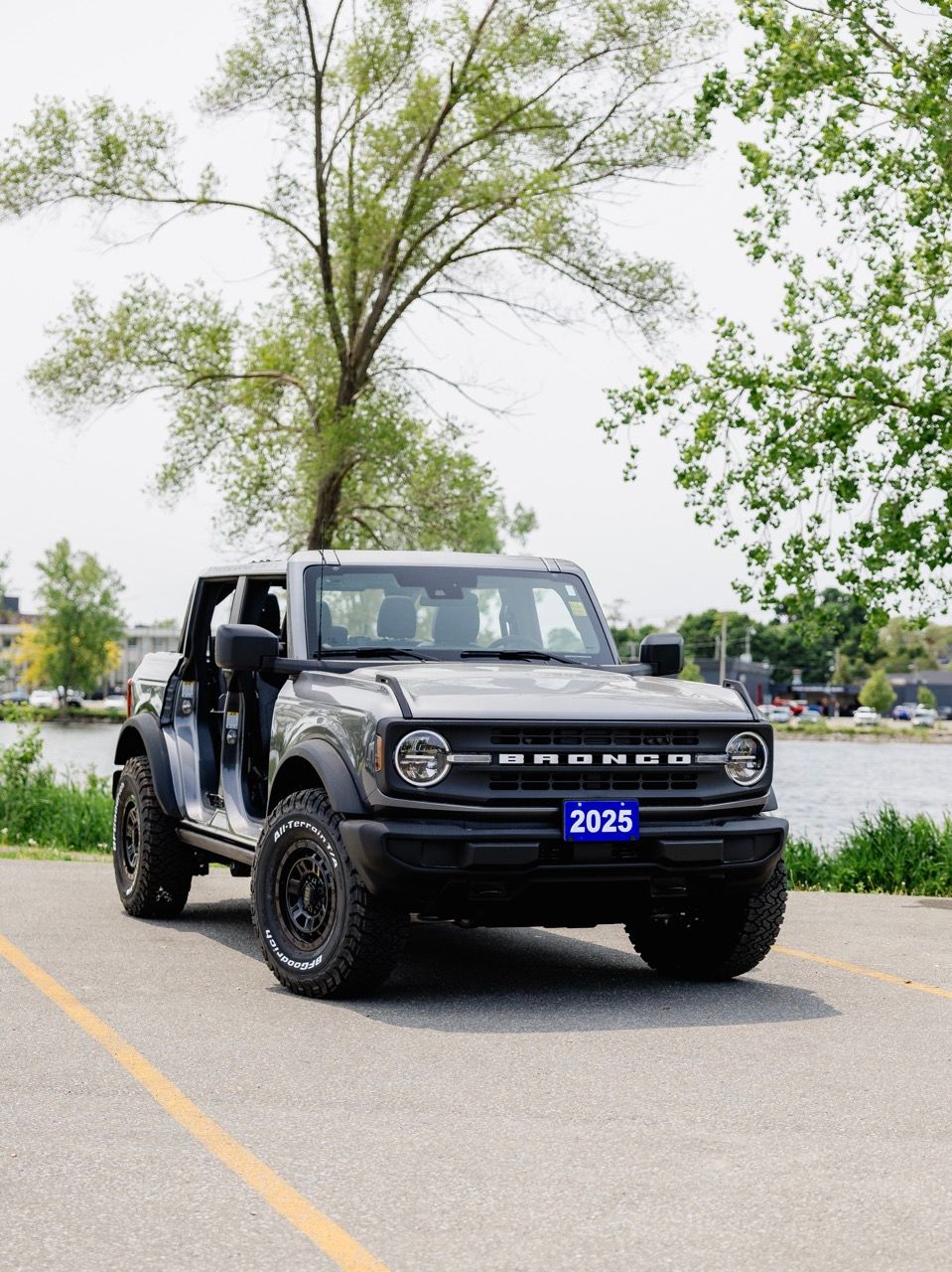 2025 Ford Bronco 4-door in dark gray with doors removed, parked near a waterfront with trees.