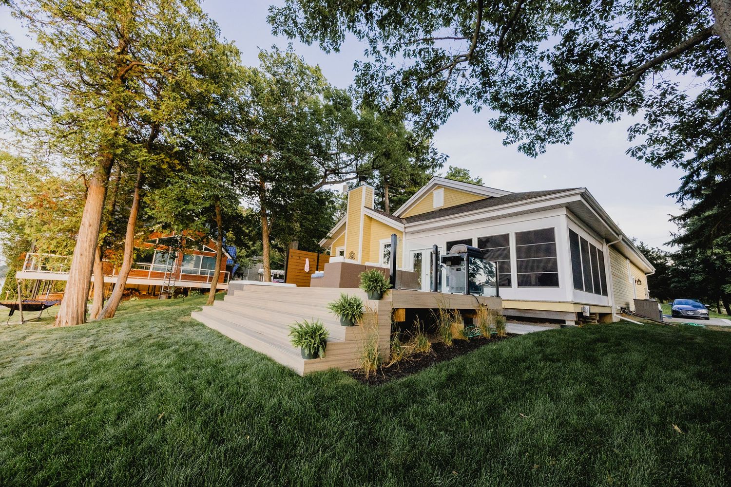 Yellow lakeside cottage with sunroom, multi-level deck, and lush green landscaped yard.
