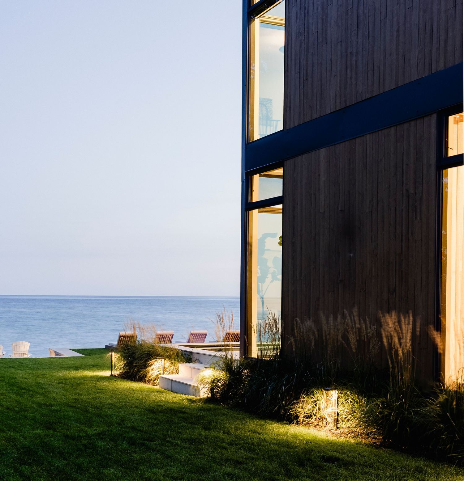 Modern dark wood exterior home at dusk with large windows, lawn, and serene ocean views.