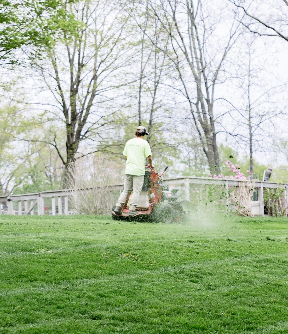 Person riding stand-on mower across green lawn surrounded by trees in spring landscape.