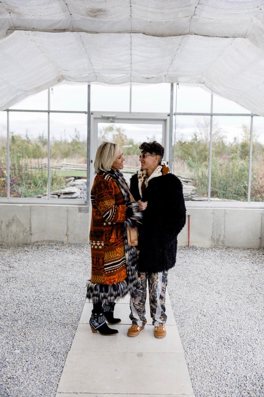 Two people in bold, layered outfits face each other inside a bright, unfinished greenhouse structure.