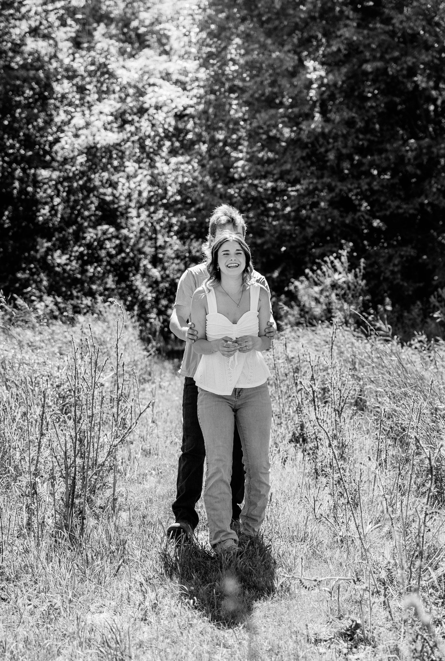 Black and white photo of a smiling couple embracing in a sunlit meadow surrounded by lush trees.