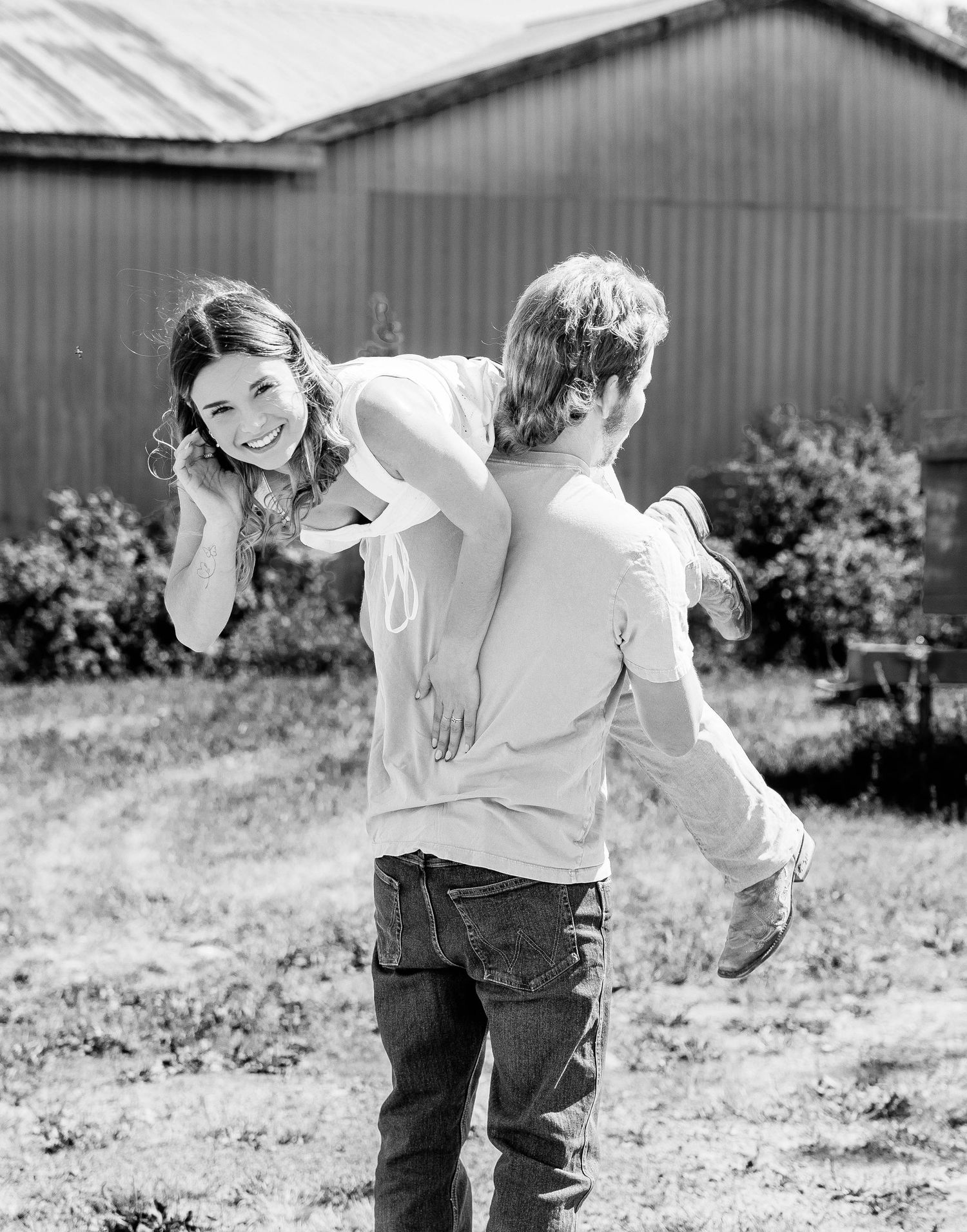 Black and white photo of a smiling woman giving a child a piggyback ride outdoors near a barn.