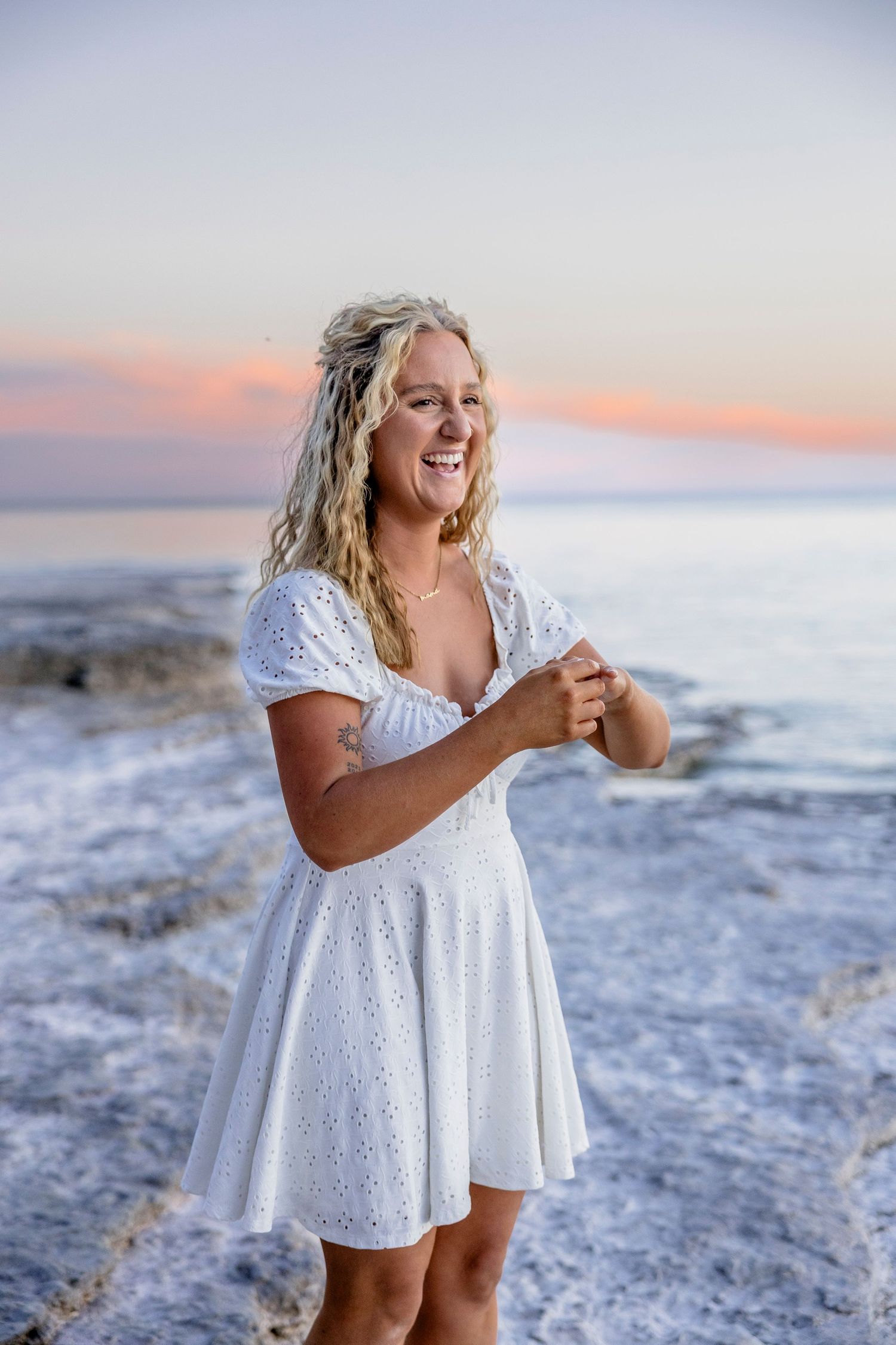 Happy blonde woman in white lace dress laughing on rocky shoreline at sunset.