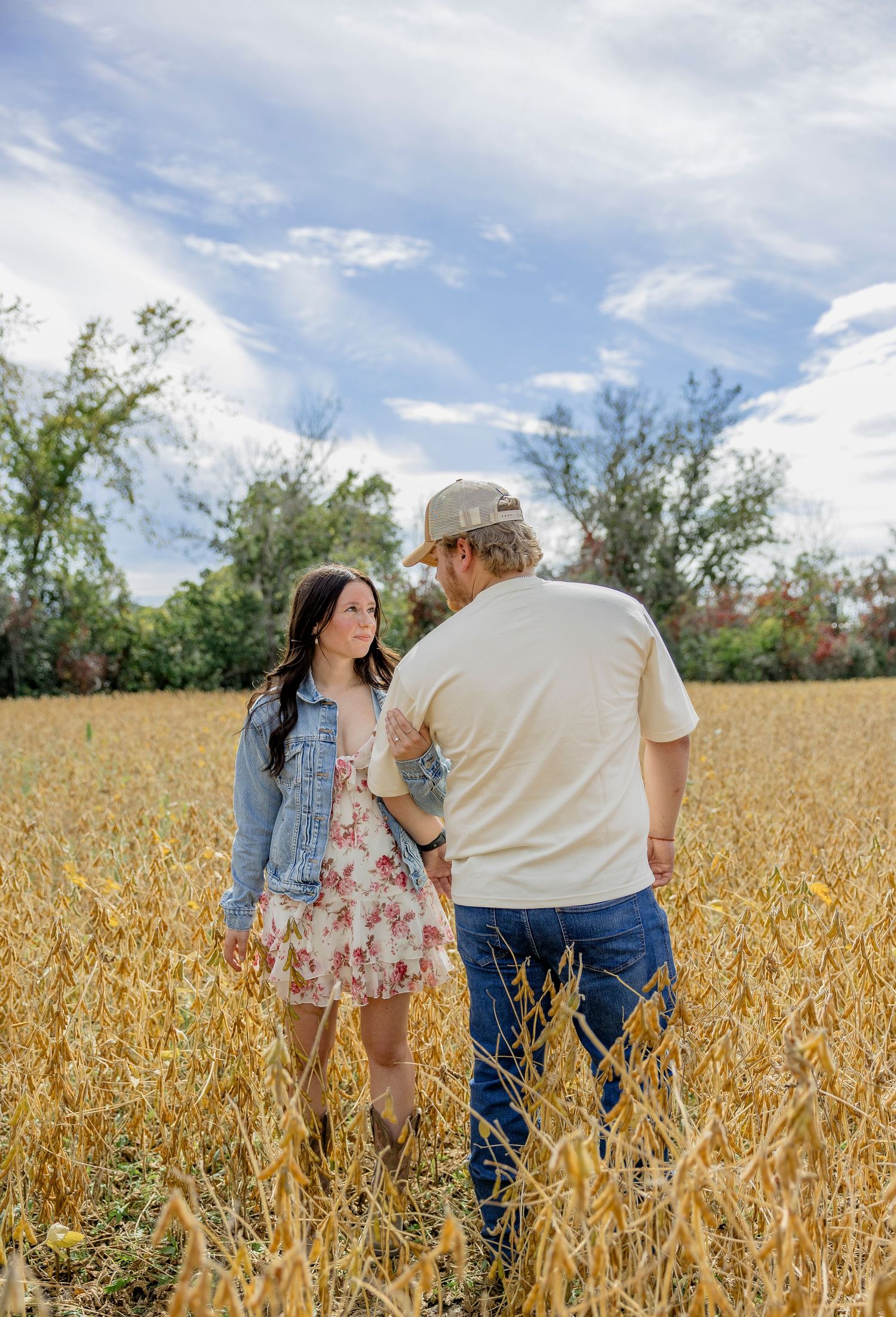 Couple standing in golden wheat field, woman in floral dress and denim jacket, man in cream shirt and cap, trees behind.