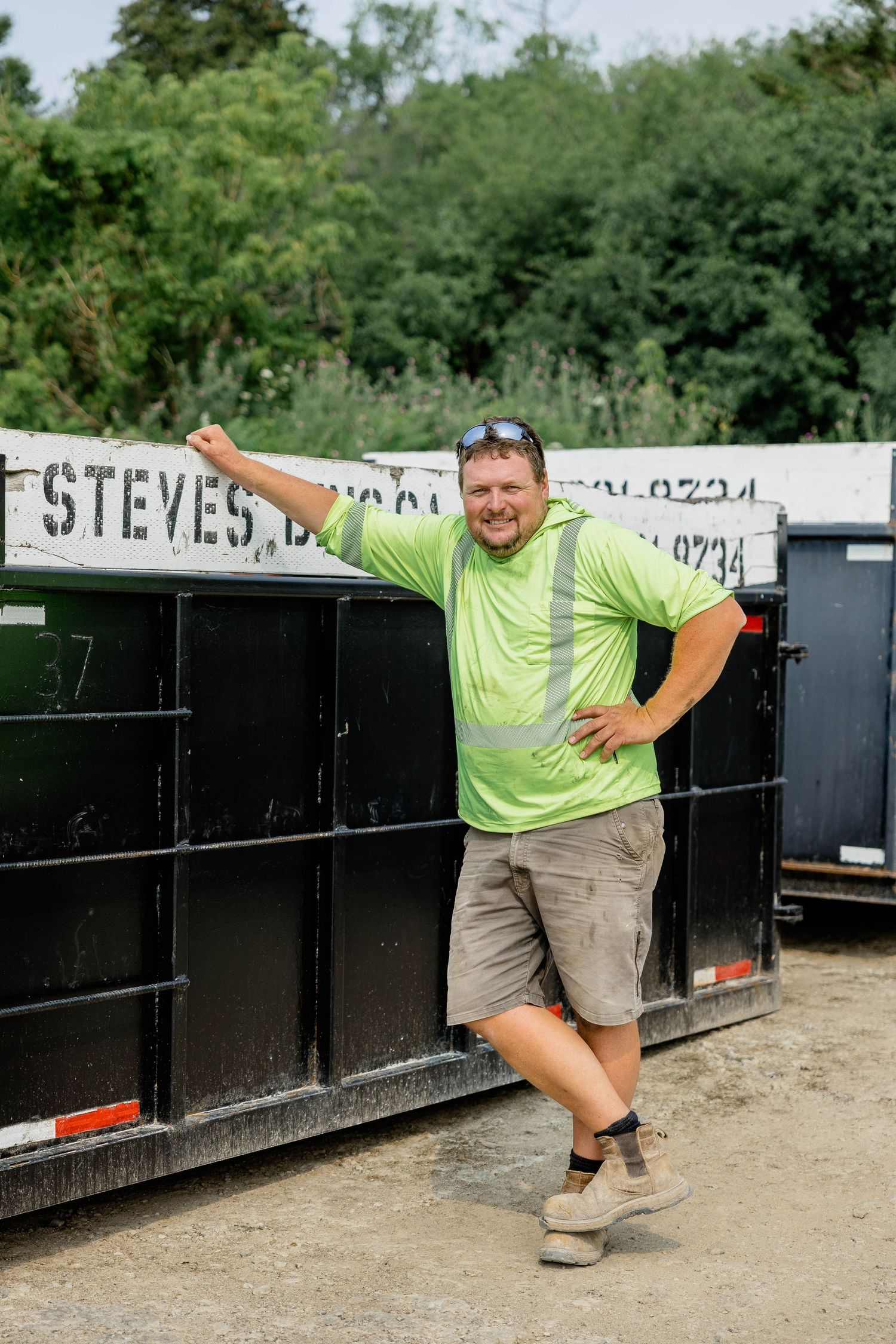 Smiling worker in green safety shirt poses proudly beside Steve's Disposal dumpsters outdoors.