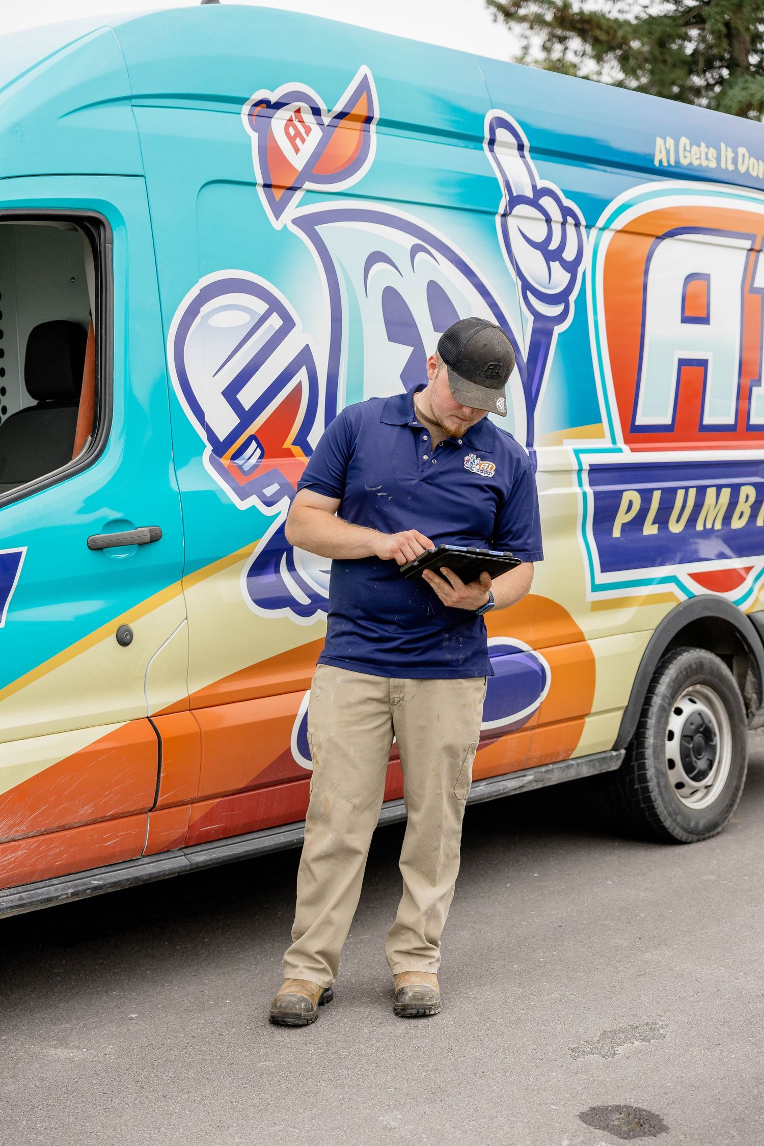 A plumber in uniform reviews a tablet outside a colorful branded service van for A1 Plumbing.