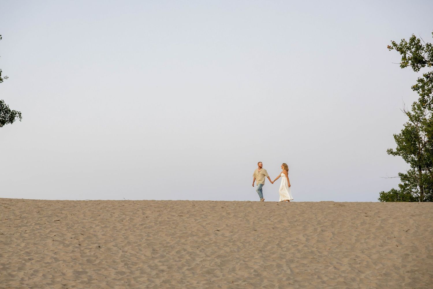 Couple holding hands on sandy dunes with trees framing the horizon under a clear sky.