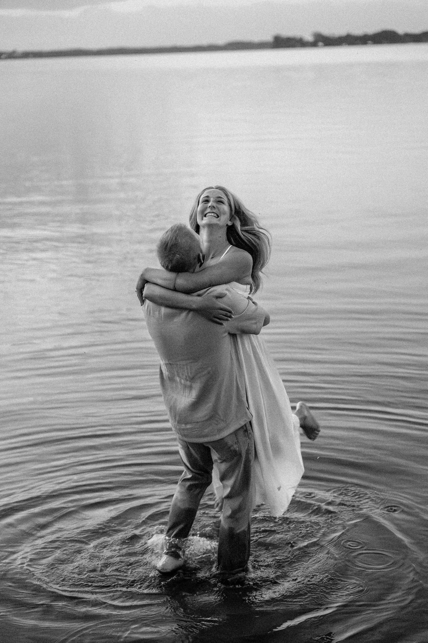 Black and white photo of a couple embracing in a lake, woman laughing joyfully as man lifts her in shallow water.