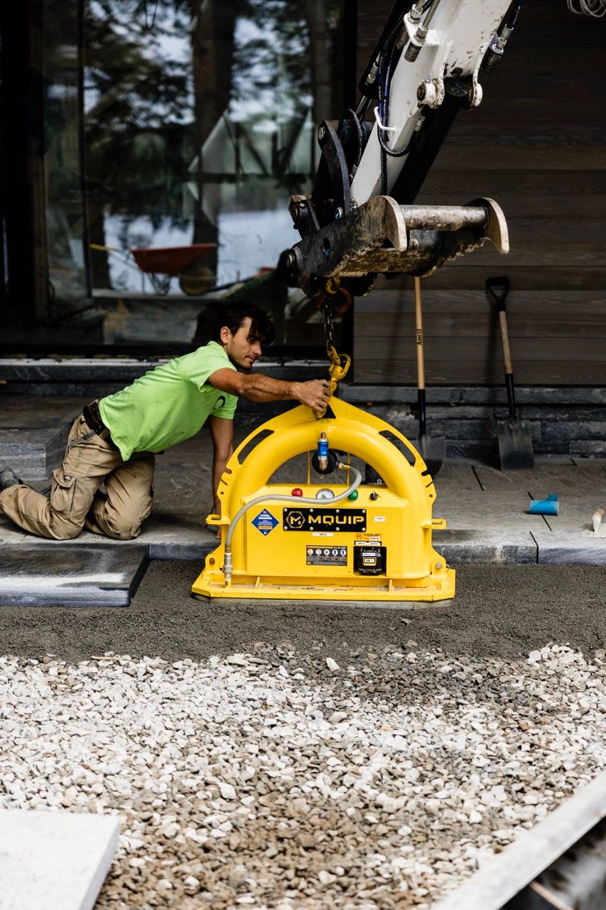 Worker in green shirt kneeling to adjust a yellow MQUIP vacuum lifter attached to an excavator arm.