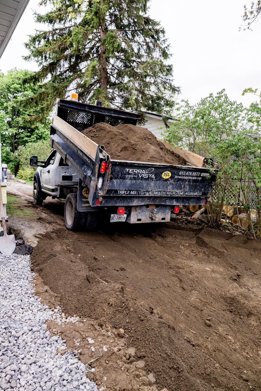 Terra Vista dump truck depositing load of soil beside white house and gravel landscaping area.