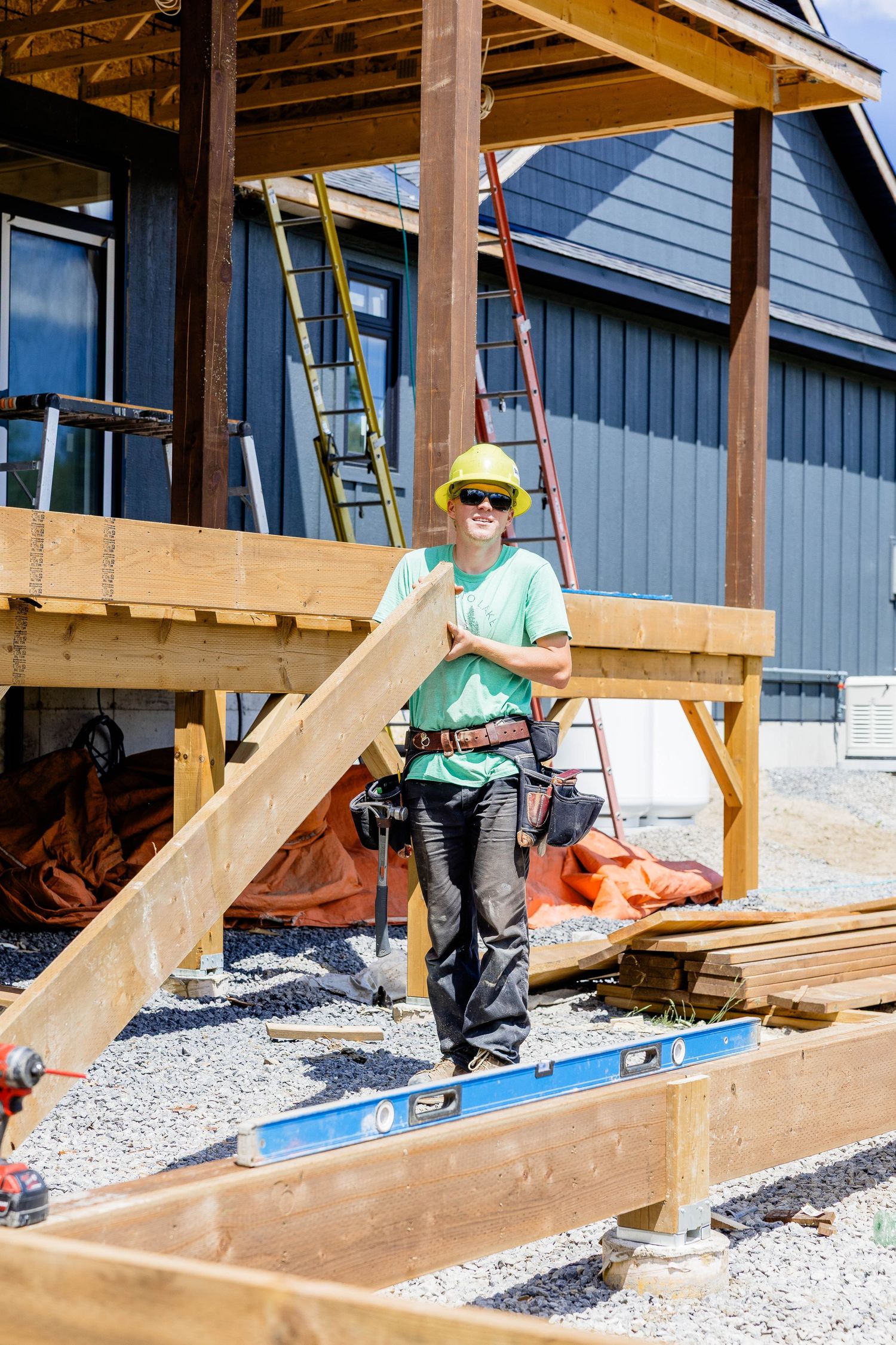 Smiling construction worker in yellow hard hat holding a wooden beam at a residential building construction site.