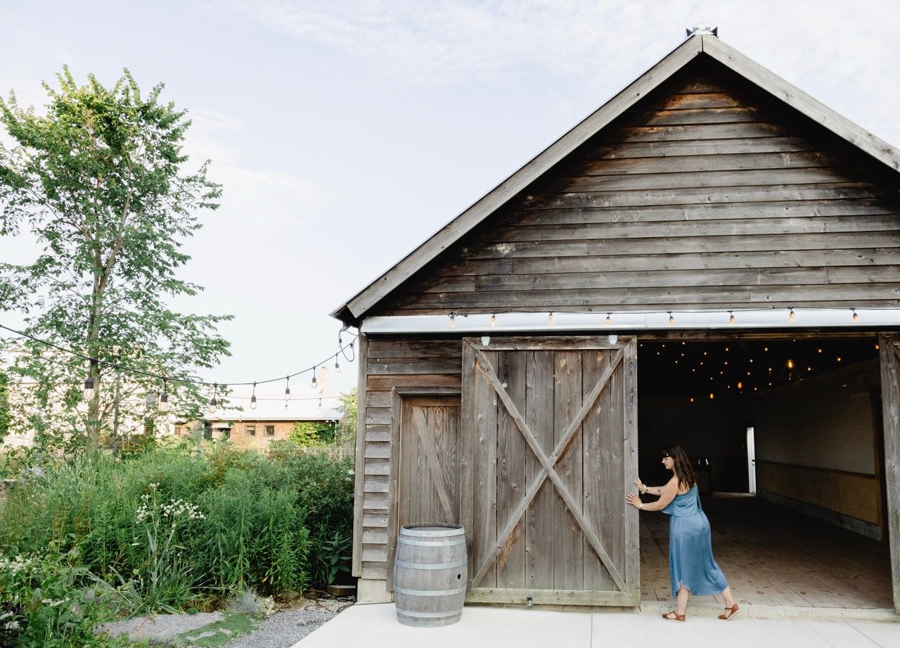 Woman in blue dress opening large wooden barn doors with string lights outside.