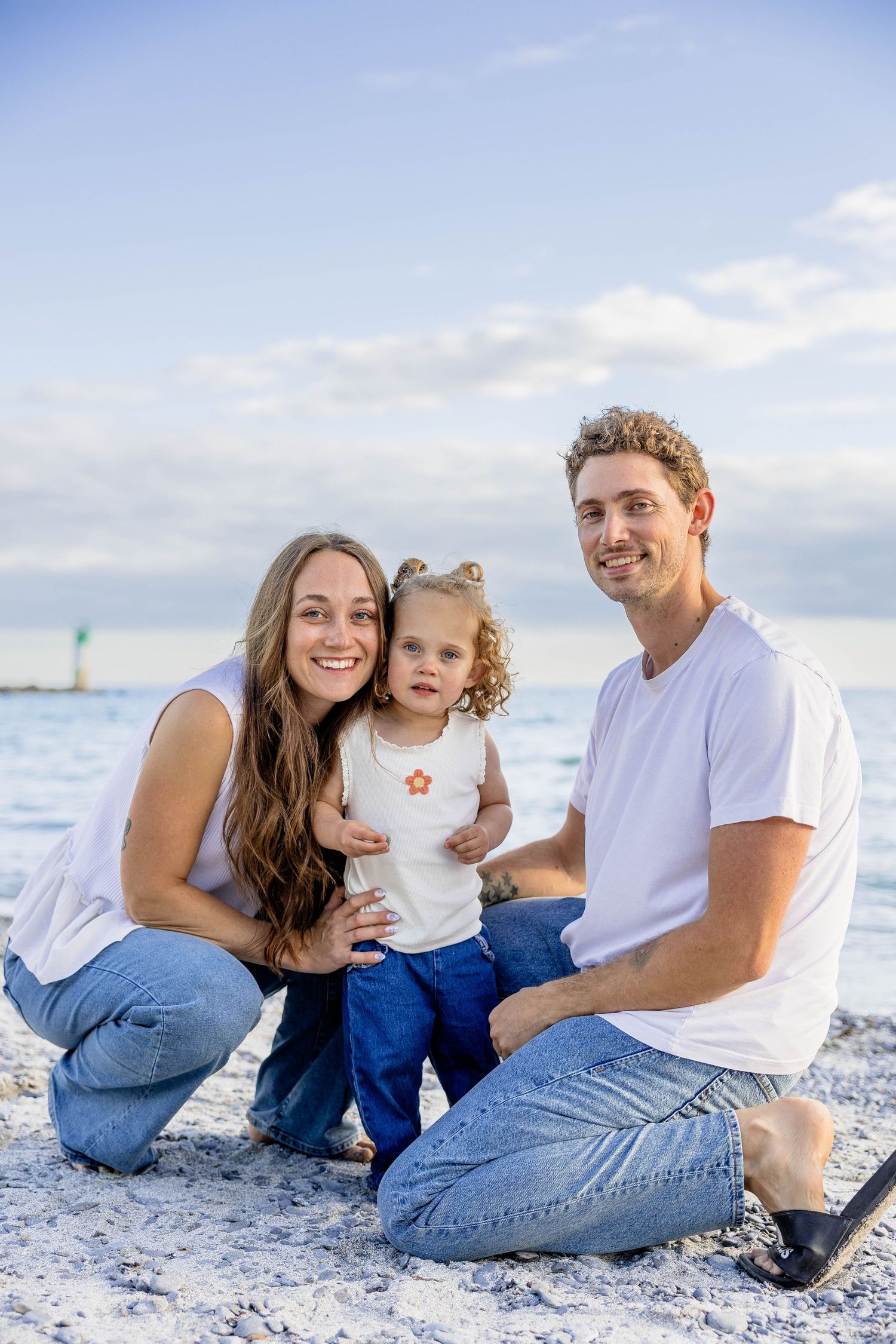Happy family of three posing on a sandy beach, parents in white shirts and jeans with their toddler daughter by the ocean.