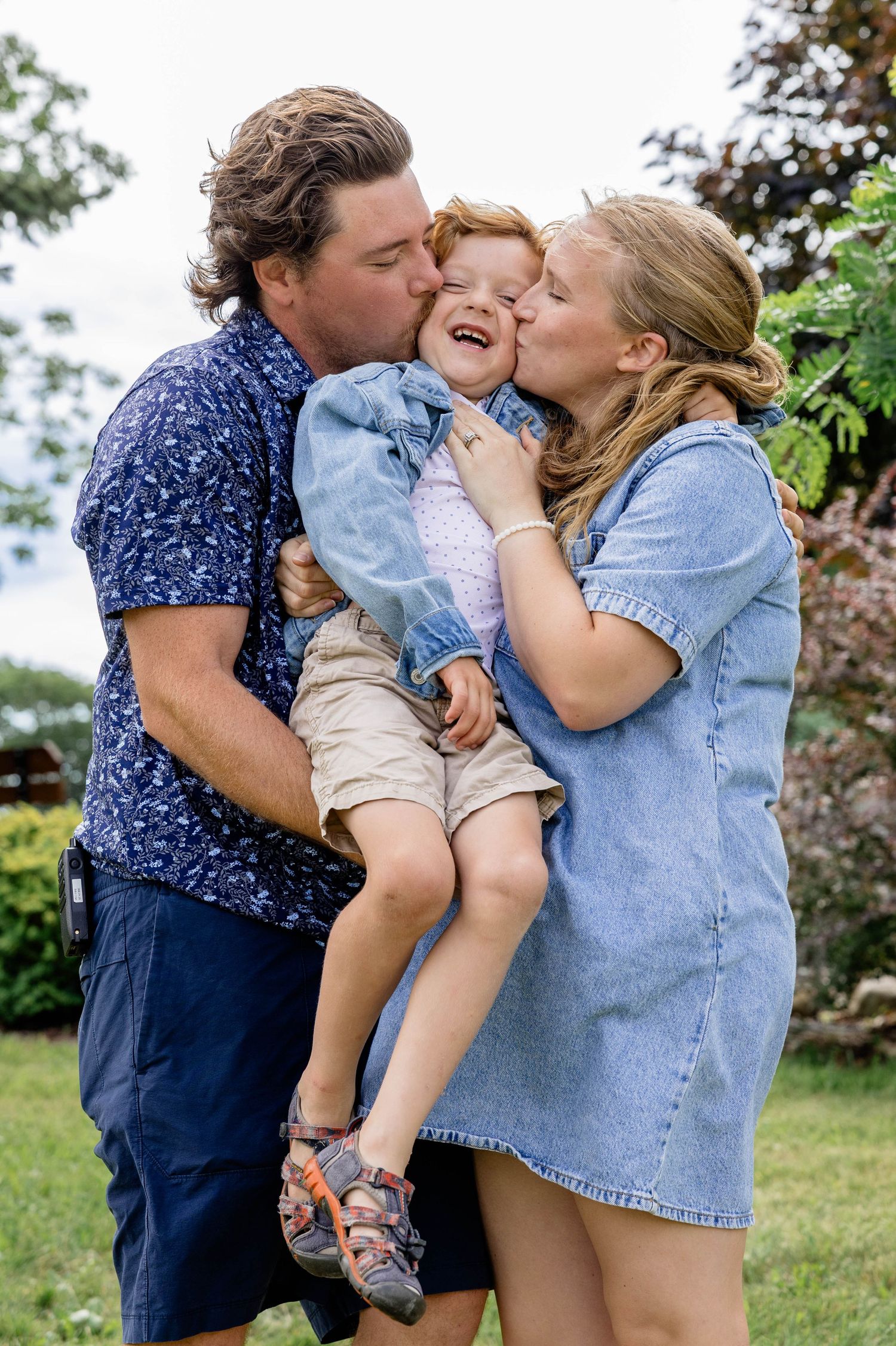 Happy family moment with parents kissing their laughing red-haired toddler outdoors in a lush green garden.