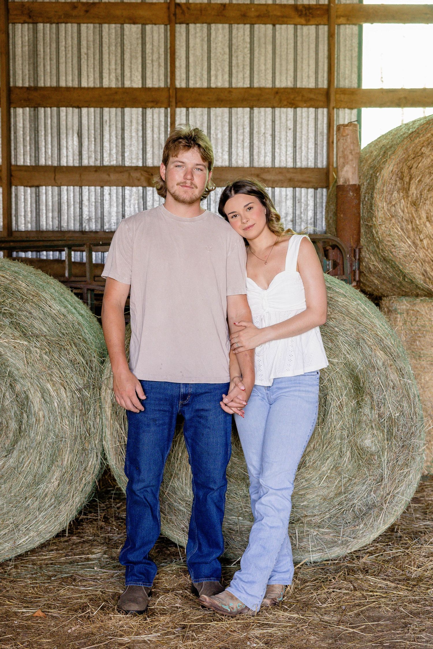 A young couple poses in front of round hay bales in a rustic barn, wearing casual jeans and simple tops.