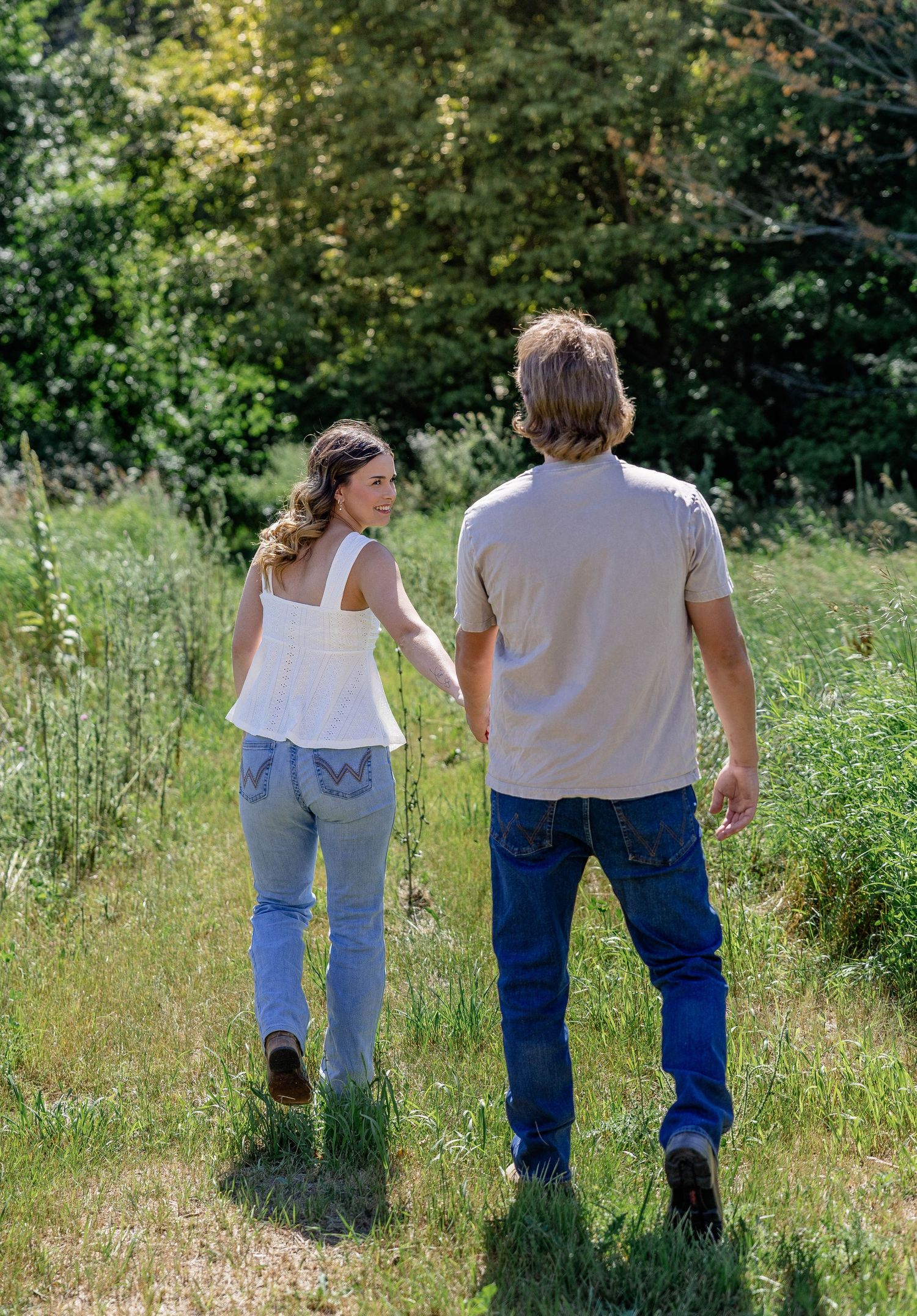 Couple holding hands walking through a lush green meadow path, woman in white top and jeans glancing back.