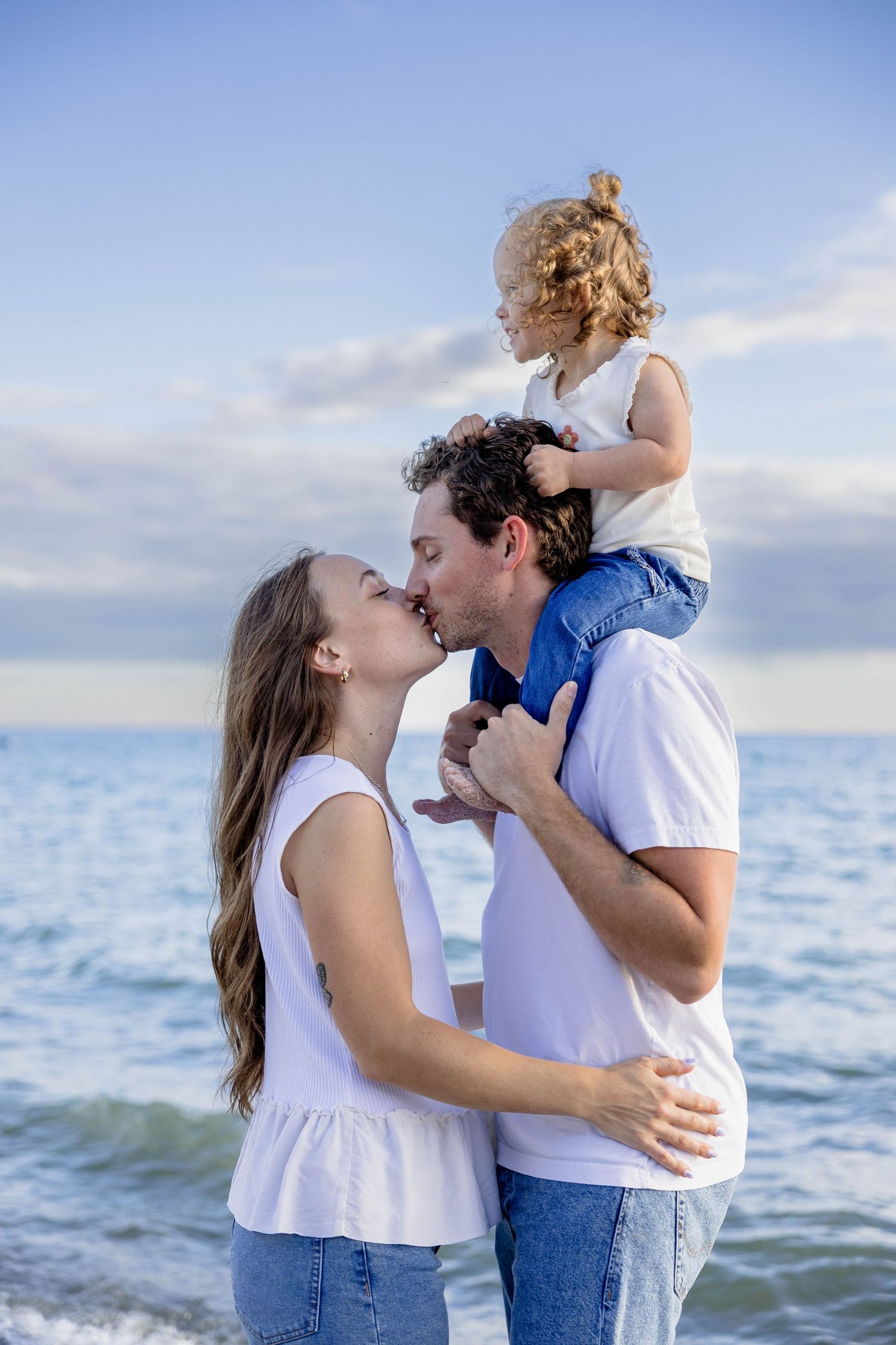 Happy young family kissing at the beach, with toddler sitting on fathers shoulders against blue sky.