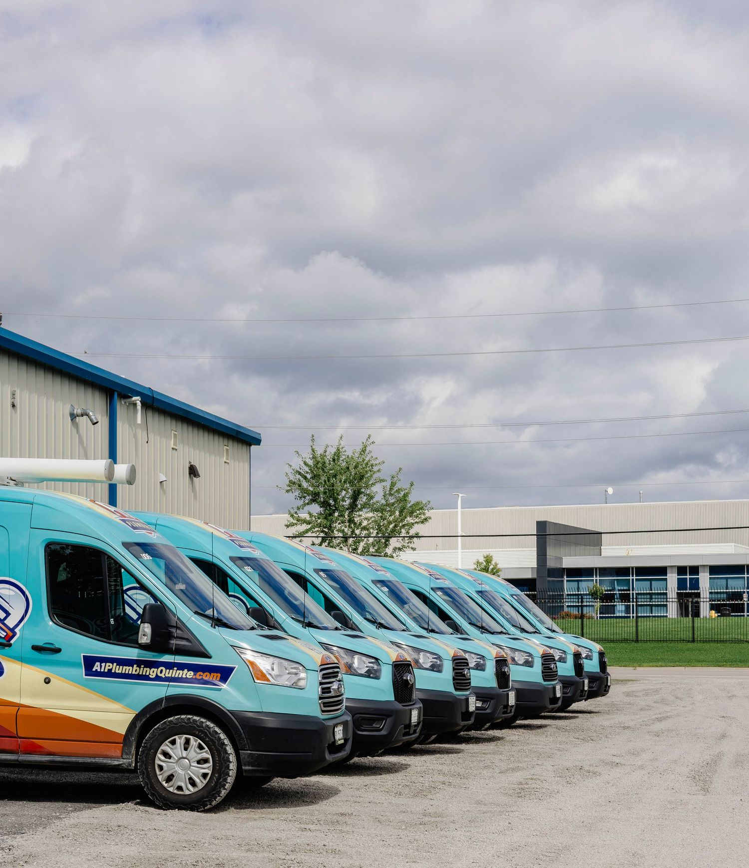 Fleet of branded teal plumbing service vans lined up in a parking lot outside a commercial building on a cloudy day.