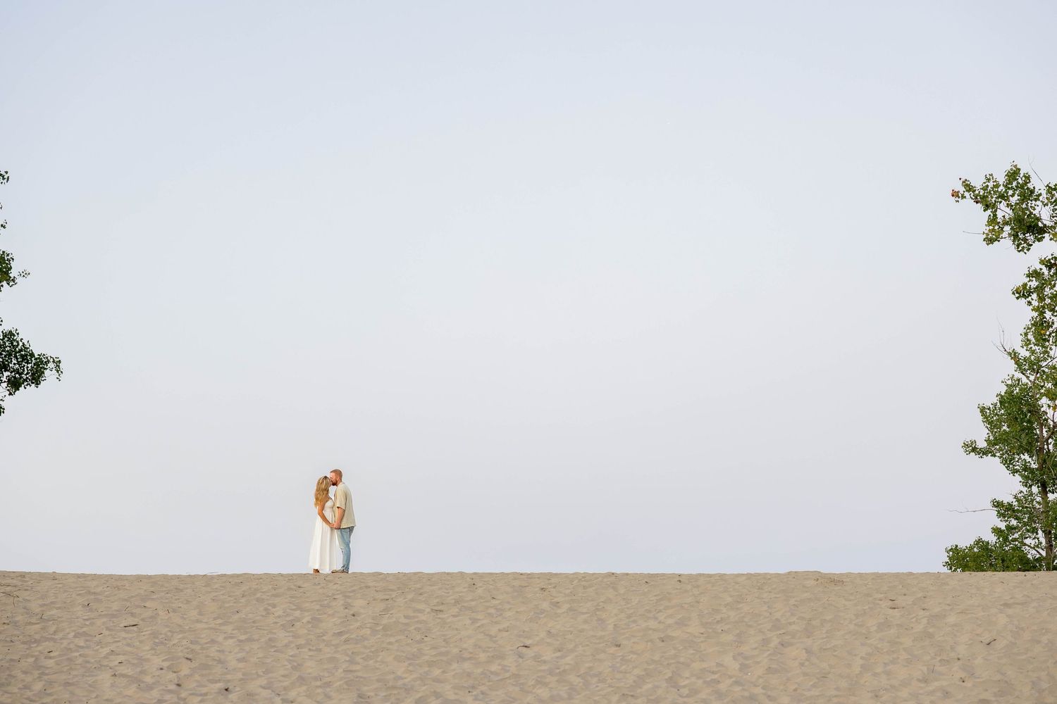 Couple embracing on sandy dunes with trees framing the scene under a clear sky.
