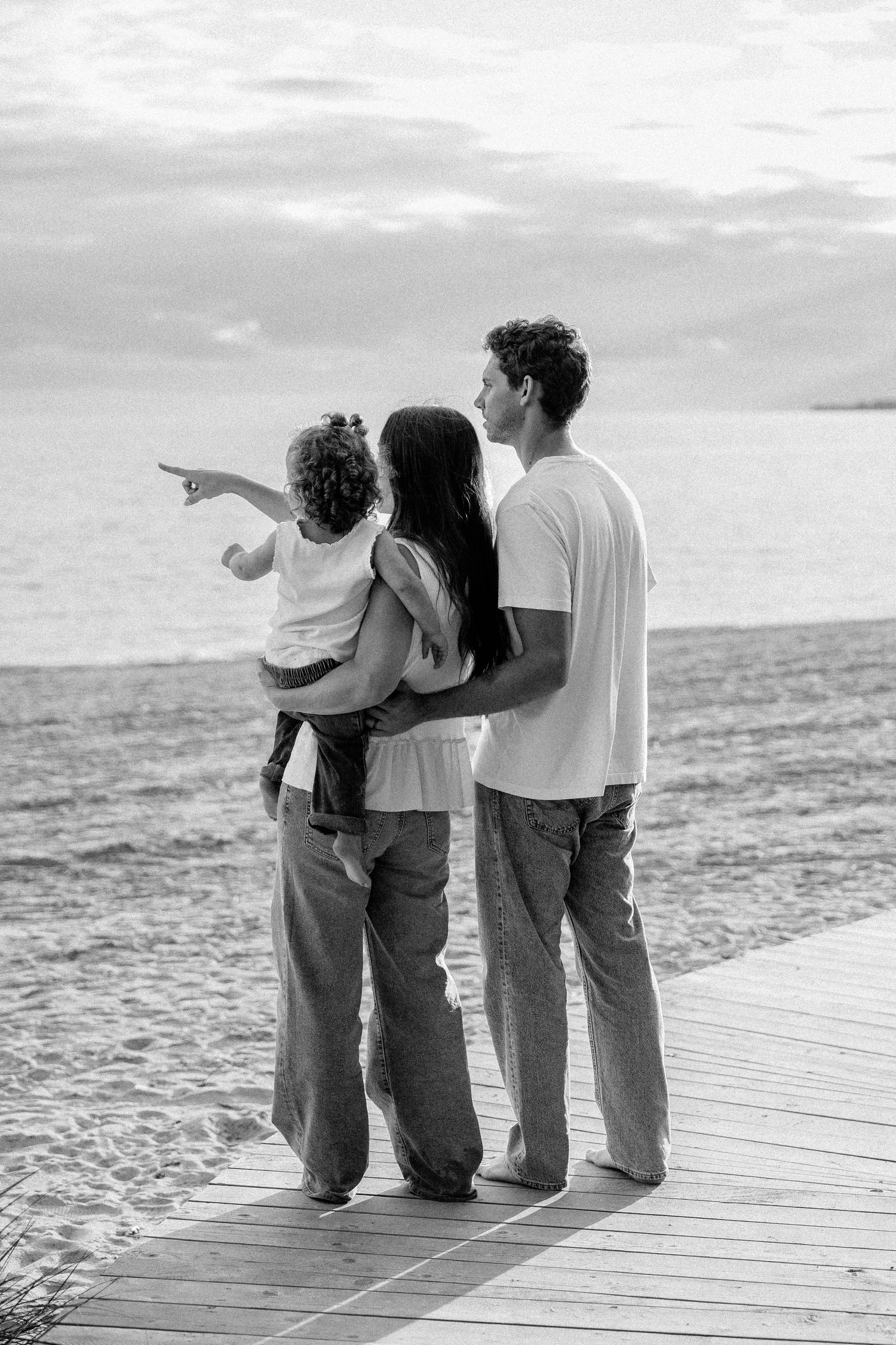 Black and white photo of a family of three standing on a boardwalk by the ocean, child pointing at the horizon.