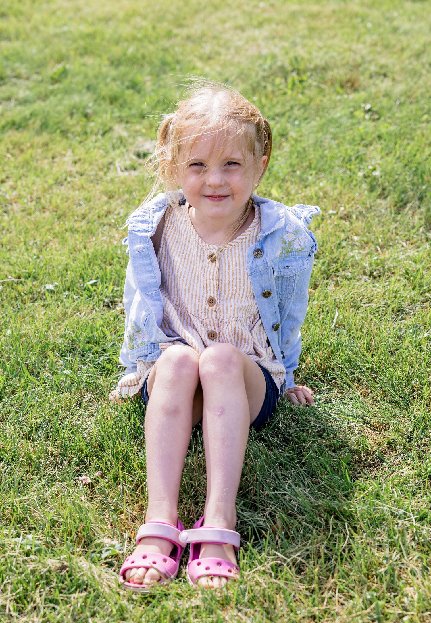 Young blonde girl in pink sandals and denim jacket sitting on green grass, smiling outdoors.
