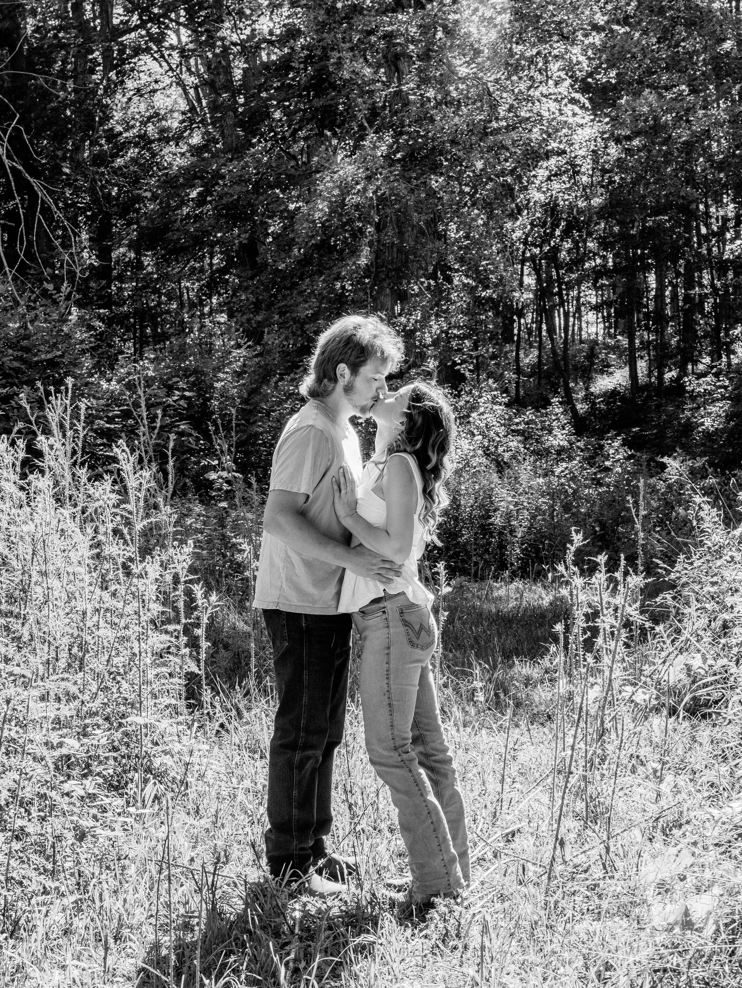 Black and white photo of a couple embracing and kissing in a wild meadow surrounded by lush trees.
