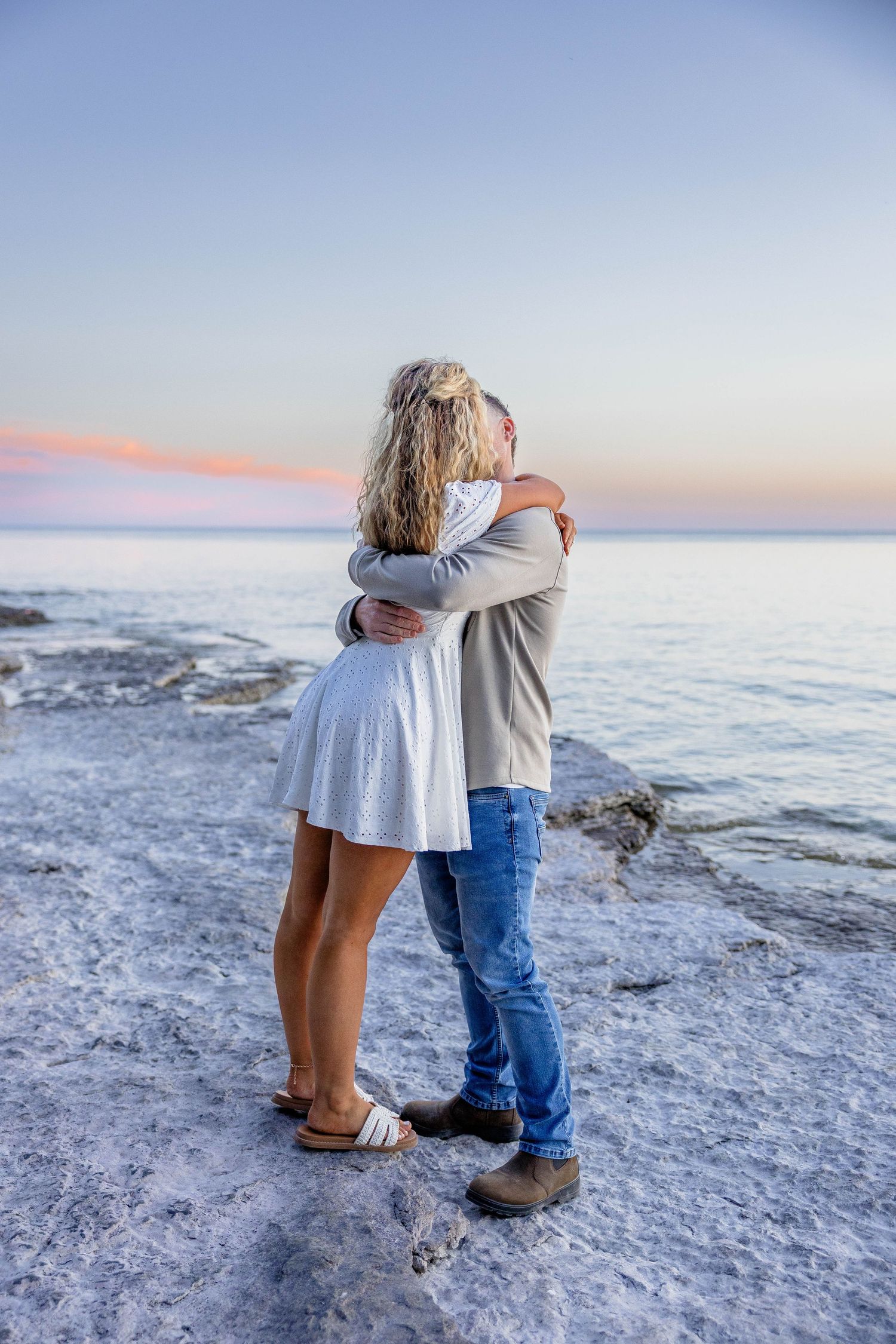 Couple embracing romantically on rocky shoreline at sunset, woman in white dress and man in jeans with ocean backdrop.