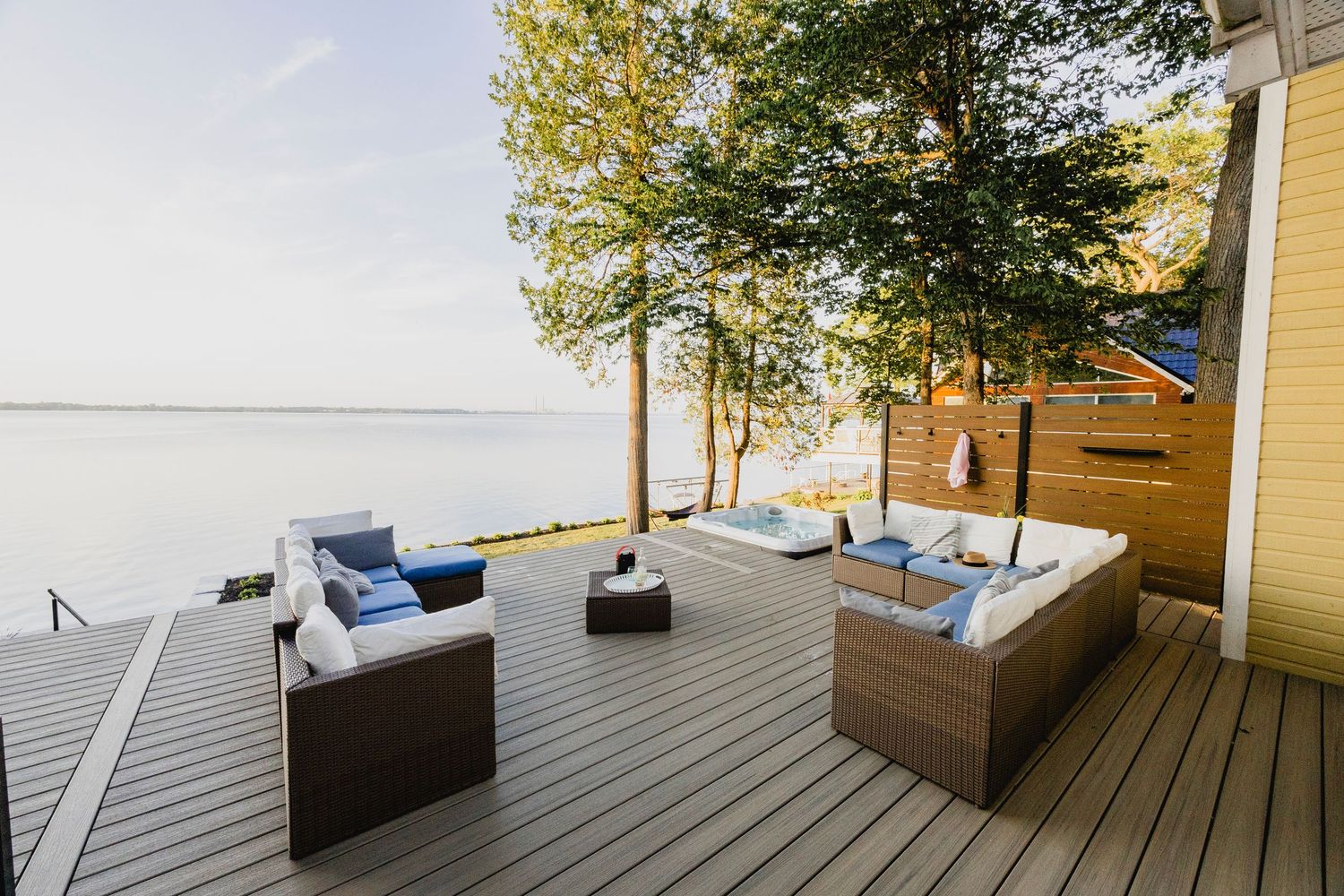 Waterfront deck with wicker patio furniture overlooking calm lake surrounded by mature trees.