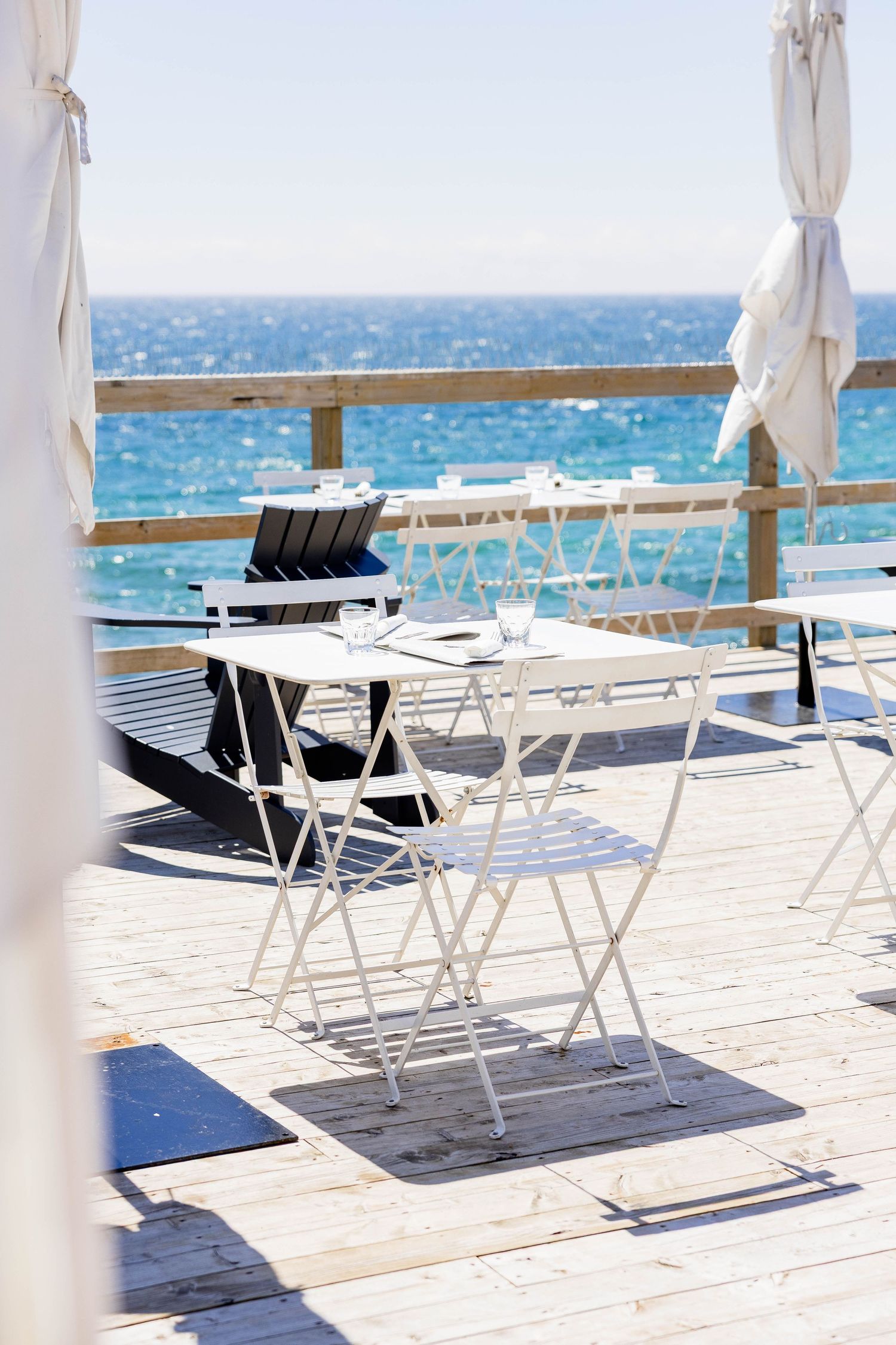 White metal chairs and tables on wooden beach deck overlooking turquoise ocean under bright sky.