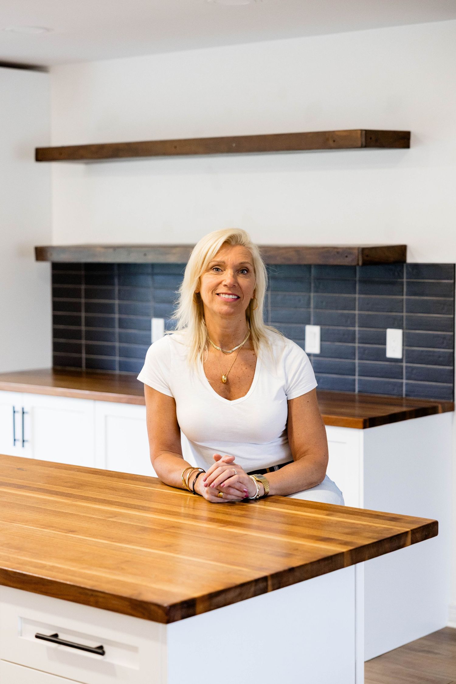 Blonde woman smiling at modern kitchen island with wood countertop, white cabinets, and blue tile backsplash.