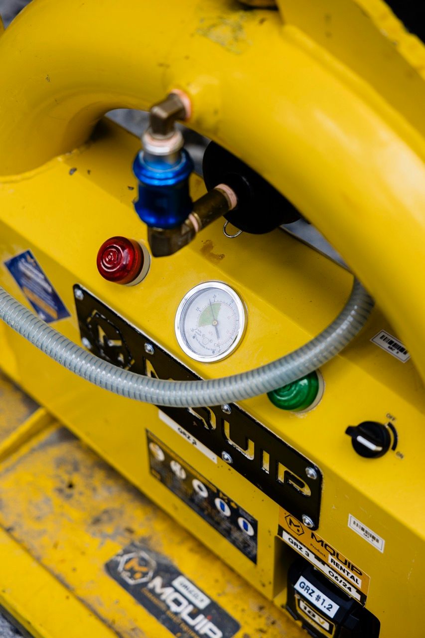 Close-up of yellow MQUIP vacuum lifter control panel showing pressure gauge, indicator lights, and hose fittings.