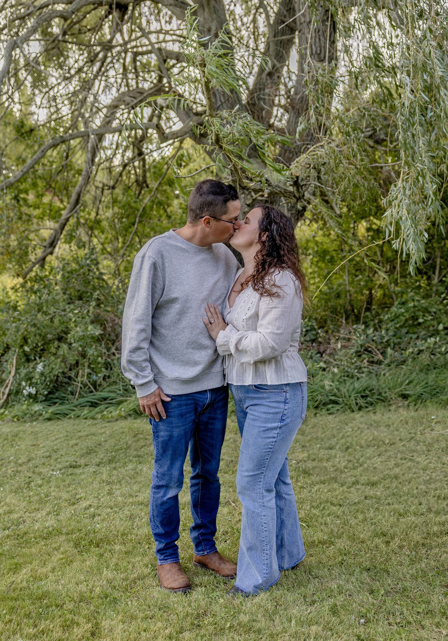 Couple kissing under a weeping willow tree on green grass, wearing casual jeans and sweaters in a lush garden setting.