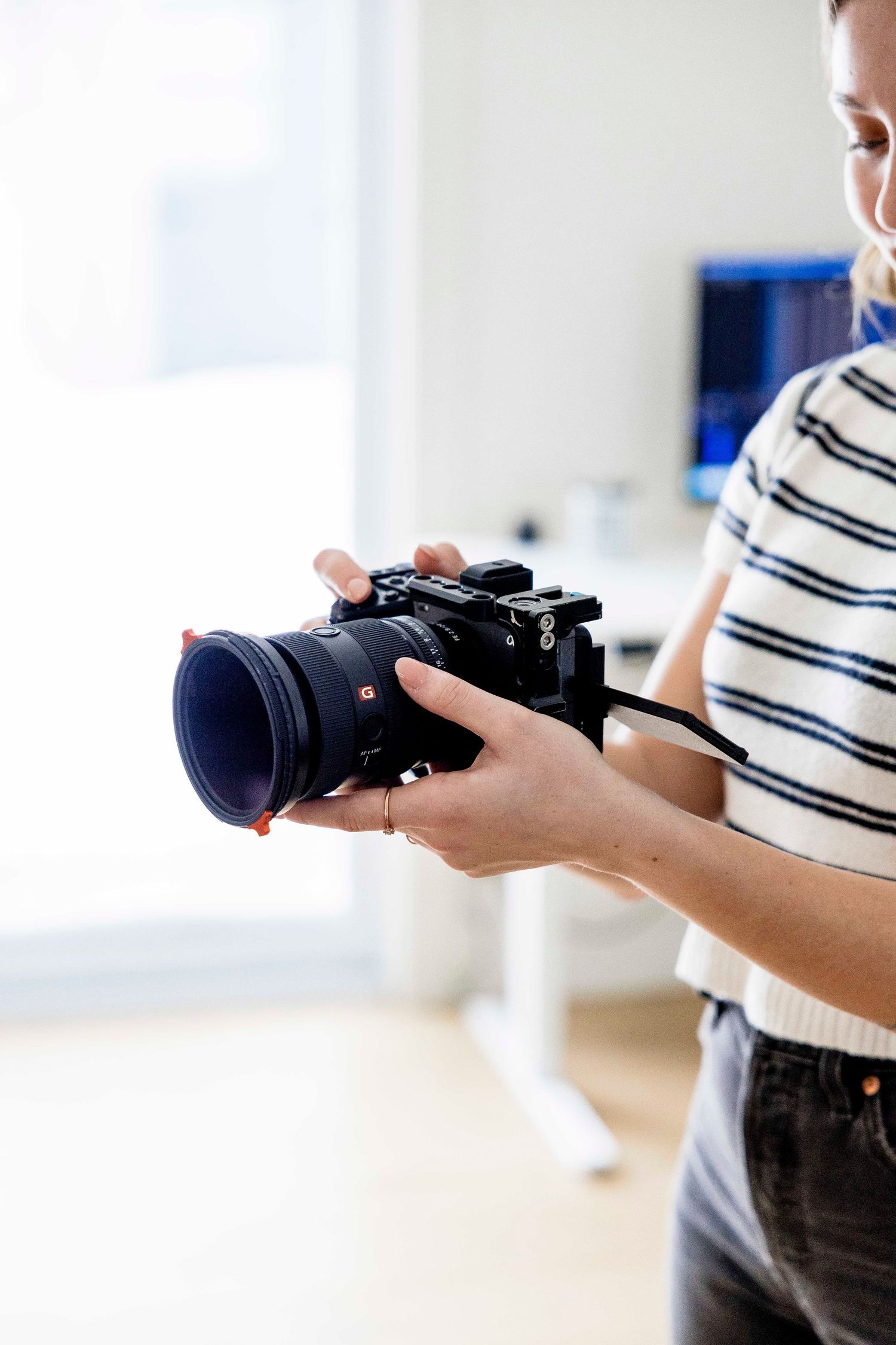 A photographer holds a professional Sony mirrorless camera with a large lens in a bright modern office setting.