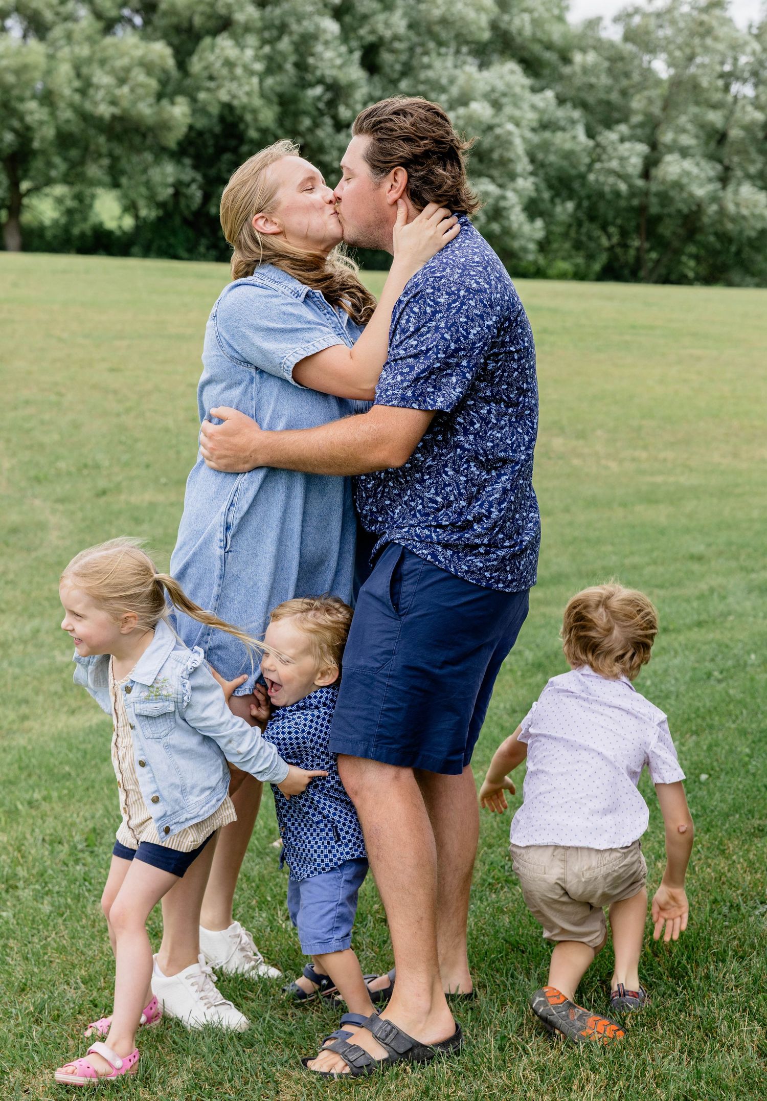Couple kissing outdoors while four young children play around them on green grass during a family photo session.