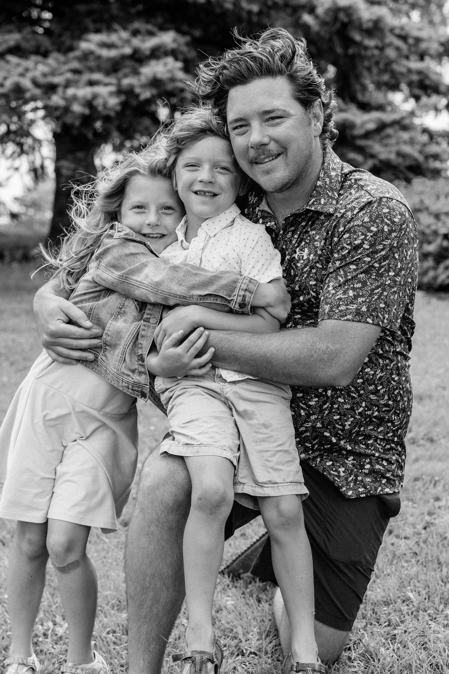 Black and white photo of a father kneeling outdoors, hugging two smiling children in a park setting.