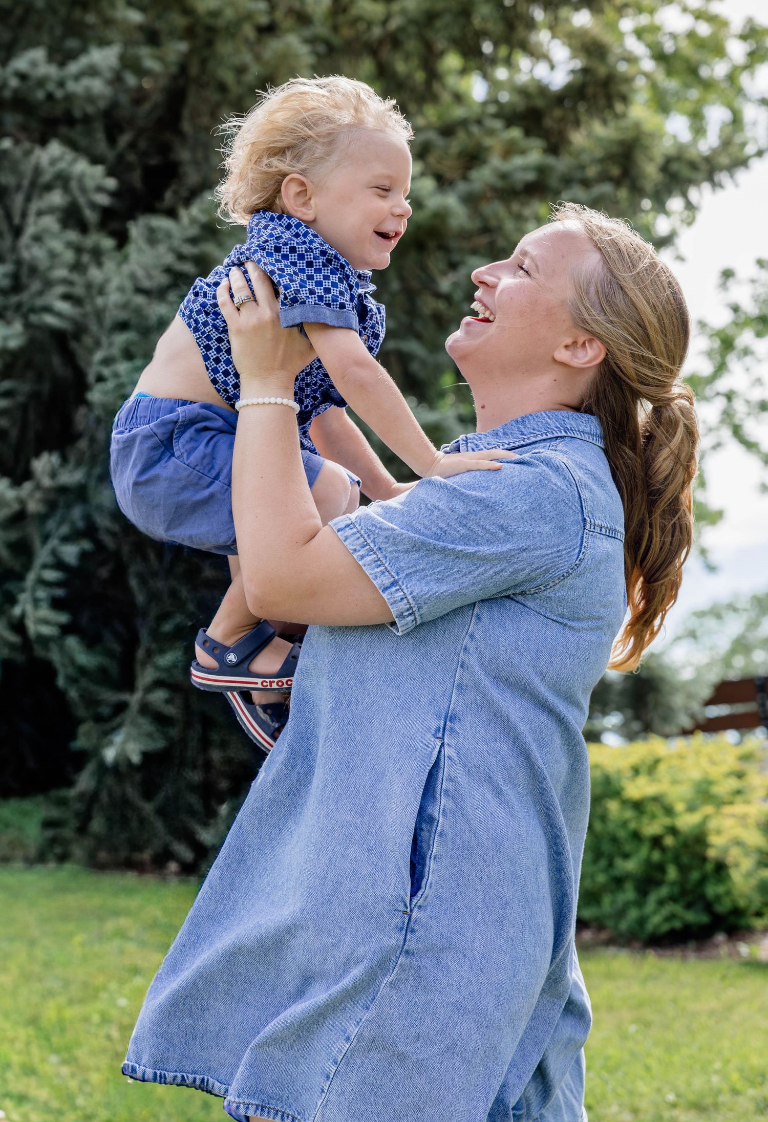 A joyful woman in denim lifts a laughing toddler in a blue dress outdoors in a lush green garden.