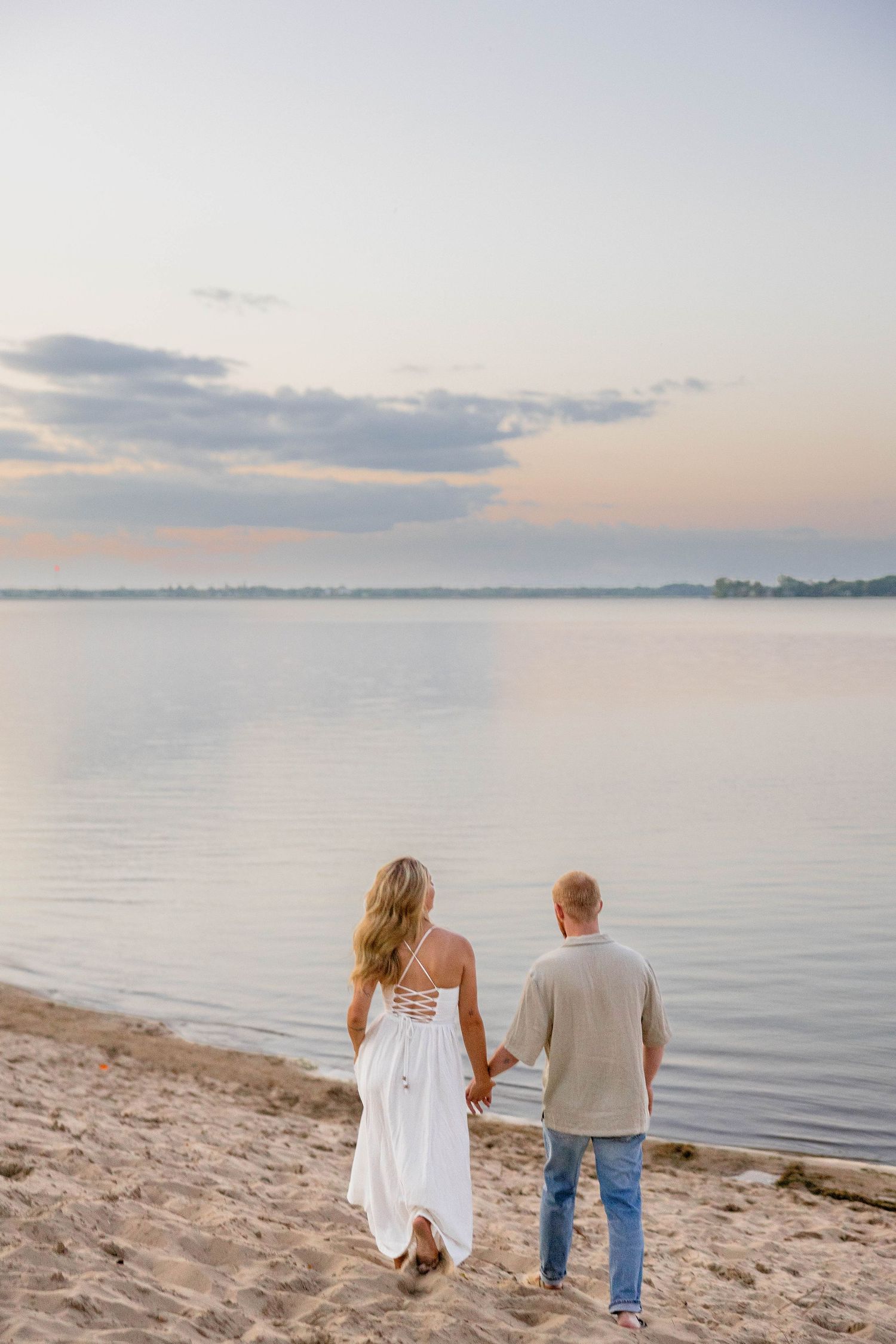Couple holding hands on sandy beach at sunset, woman in white dress and man in jeans facing calm water.
