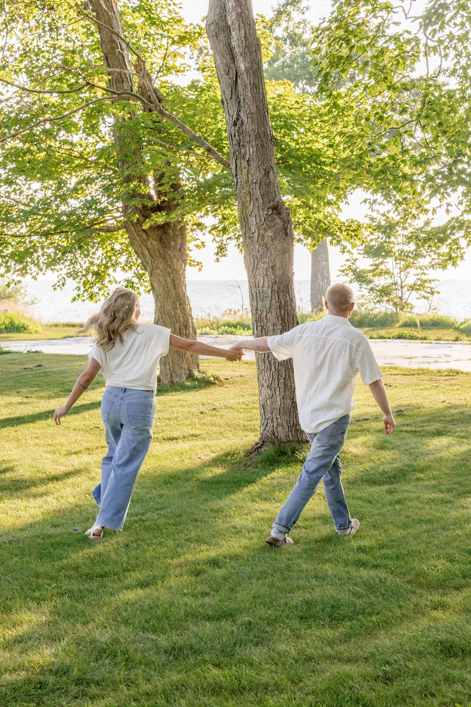 Couple holding hands walking toward trees in a sunny green park near water, both wearing white shirts and blue jeans.