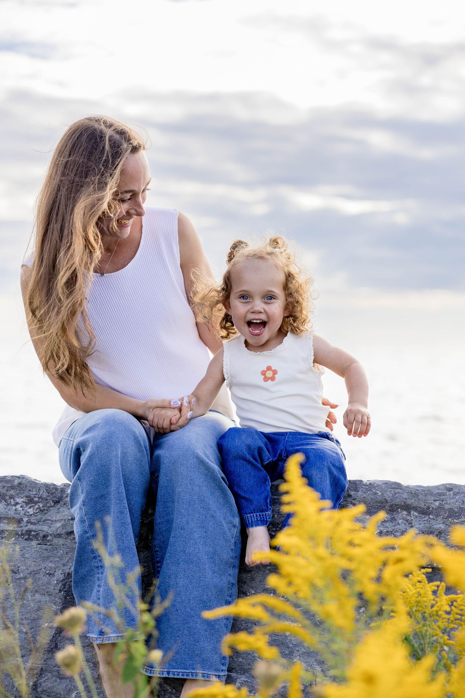 A smiling mother and her curly-haired toddler sit on rocks near the ocean, both wearing white tops and blue jeans.